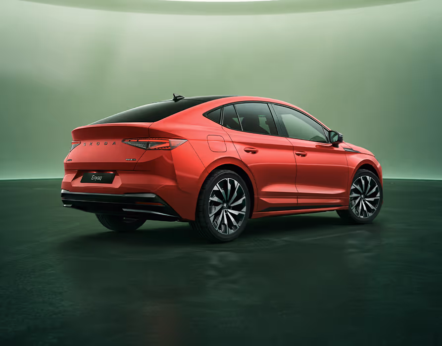 Ein rotes Škoda Enyaq Coupé Sportline steht in einer Dreiviertel-Heckansicht vor einem grünen Studiohintergrund. Das Fahrzeug hat eine abfallende Dachlinie, die in einem integrierten Spoiler endet, und ein kontrastierendes schwarzes Dach mit einer Haifischflossen-Antenne. Die Rückleuchten und der schwarze Škoda Schriftzug am Heck sind zu erkennen.