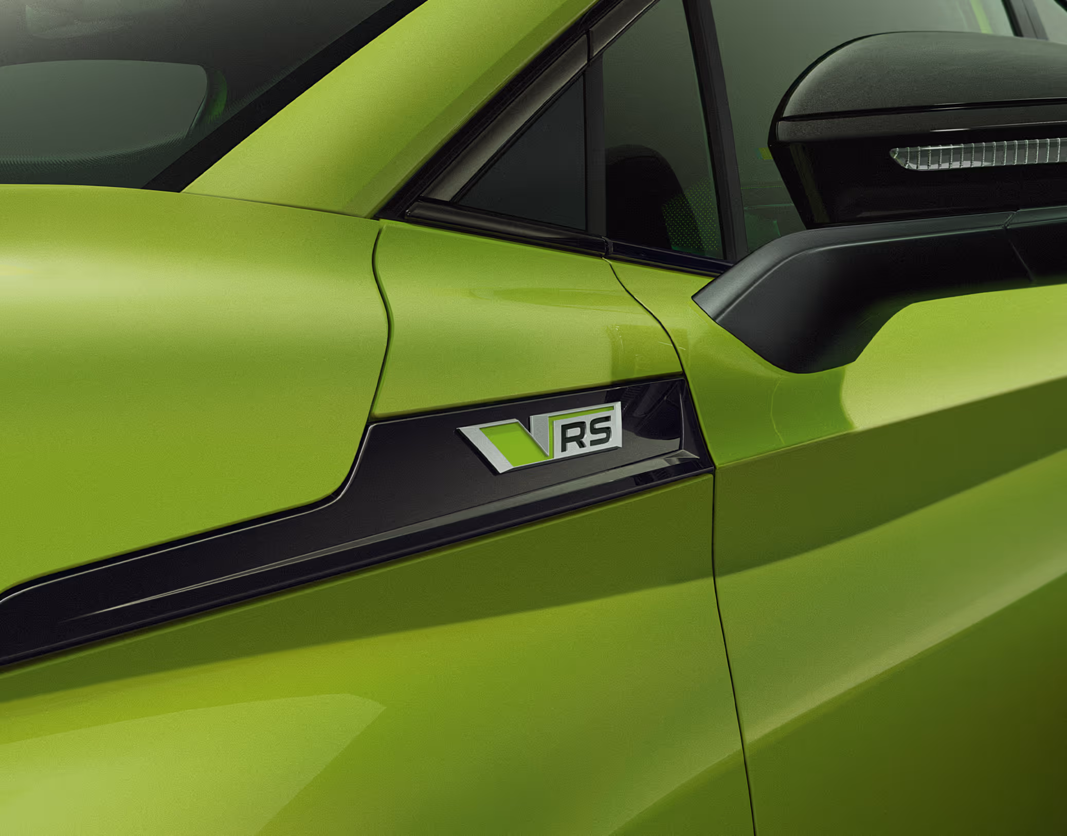 Detaillierte Nahaufnahme der Seite eines Škoda Enyaq RS in der Lackierung Mamba-Grün. Im Mittelpunkt steht die markante RS-Plakette, die auf einem schwarzen Designelement am vorderen Kotflügel angebracht ist. Die schwarz lackierte Außenspiegelkappe mit integriertem LED-Blinker setzt einen dynamischen Kontrast und unterstreicht das exklusive Design.