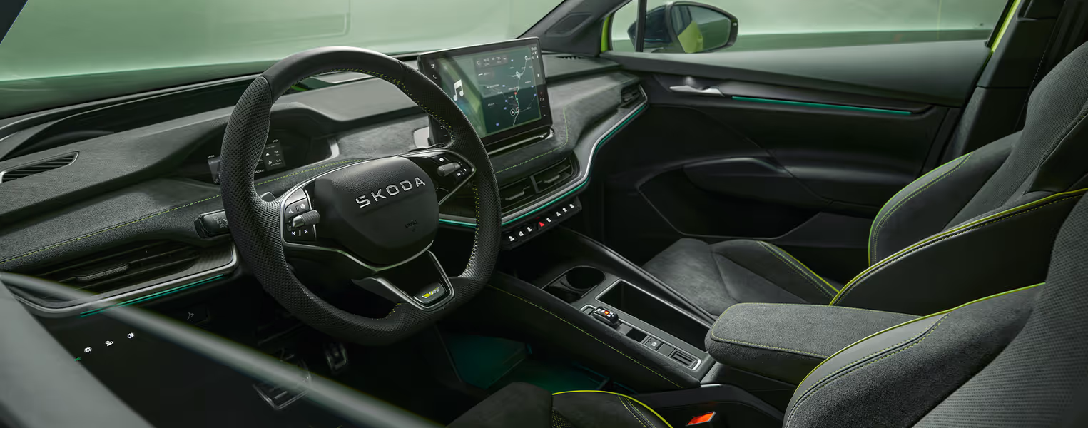 Blick durch das heruntergefahrene Seitenfenster auf der Fahrerseite ins Innere des Škoda Enyaq RS. Das sportliche Ambiente wird durch schwarze Sportsitze aus Suedia-Mikrofaser, Dekorleisten in Carbon-Optik und limettenfarbene Kontrastnähte an Sitzen, Armaturenbrett sowie dem Sport-Multifunktionslenkrad geprägt.