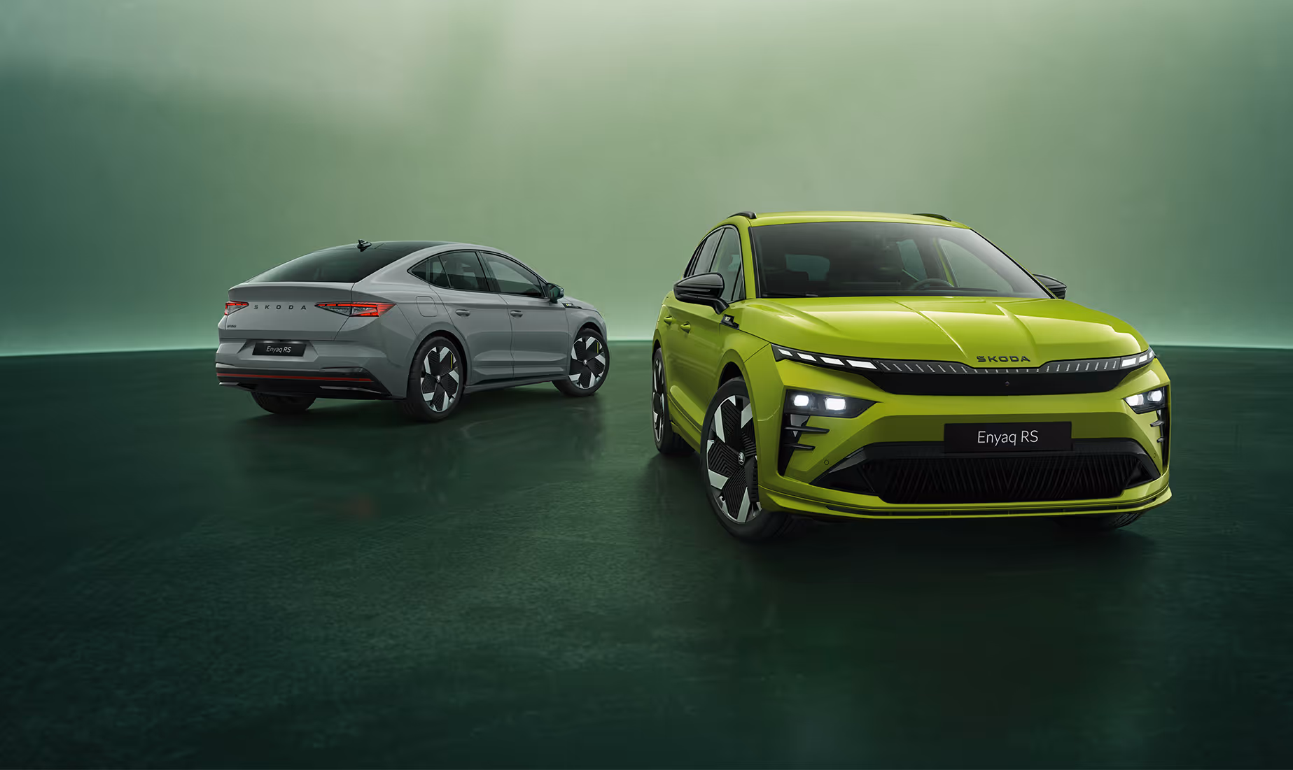 Grünes Studio-Setting. Im Vordergrund steht rechts der Škoda Enyaq RS als SUV-Variante in der Farbe Mamba-Grün in der Frontansicht. Dahinter befindet sich links das Enyaq Coupé RS in der Farbe Stahl-Grau mit seiner eleganten, abfallenden Dachlinie in der Heckansicht.