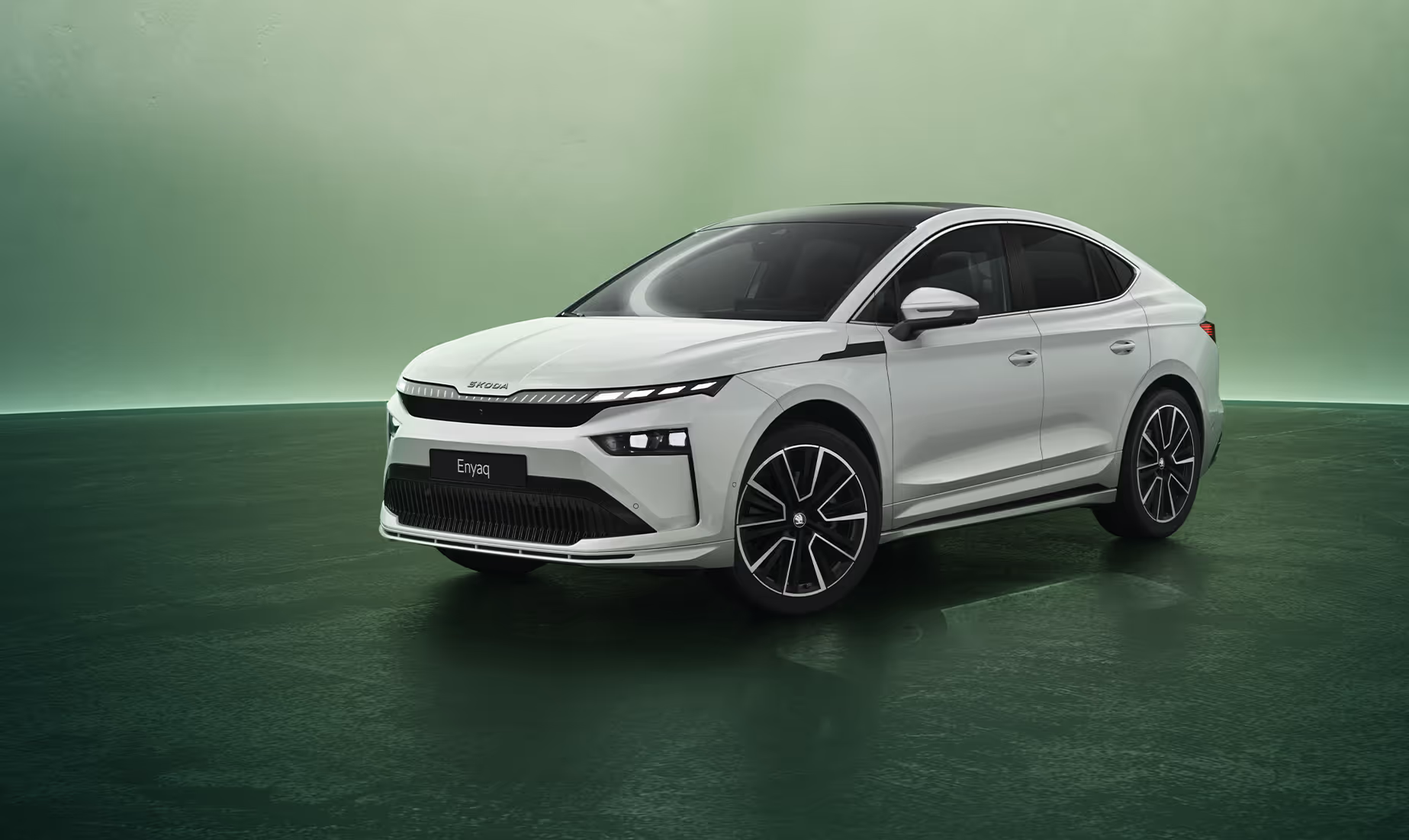 Dreiviertel-Frontansicht auf ein Škoda Enyaq Coupé in der Farbe Moon-Weiß Perleffekt vor grünen Studiohintergrund. Das elegante Design des Fahrzeugs wird durch die abfallende Dachlinie, das kontrastierende schwarze Dach und das beleuchtete Crystal Face betont. Unten rechts überlagert eine dunkelgrüne Fläche das Bild, darauf zu lesen ist der Schriftzug "Individuelle Ausstattung".