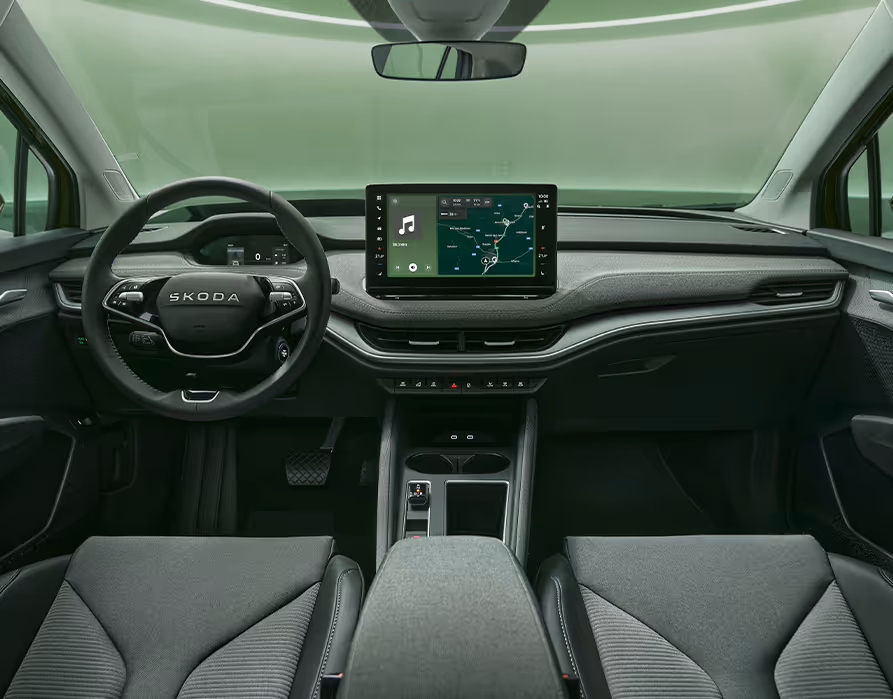 Eine symmetrische Weitwinkelaufnahme zeigt das Cockpit des Škoda Enyaq aus der Perspektive der Rücksitze. Das Interieur der Design Selection „Lodge“ zeichnet sich durch dunkle Materialien, eine durchgehende grüne Ambientebeleuchtung und das zentrale, freistehende 13-Zoll-Infotainmentdisplay aus.
