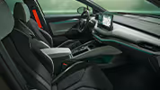Ein Blick in den vorderen Innenraum des Škoda Enyaq mit Fokus auf den Fahrer- und Beifahrersitz. Das Cockpit in der Design Selection "Lodge" ist in einer Kombination aus hellen und dunklen Materialien gehalten und wird durch eine grüne Ambientebeleuchtung akzentuiert. Ein auffälliger roter Sicherheitsgurt an der B-Säule sorgt für einen besonderen farblichen Kontrast.
