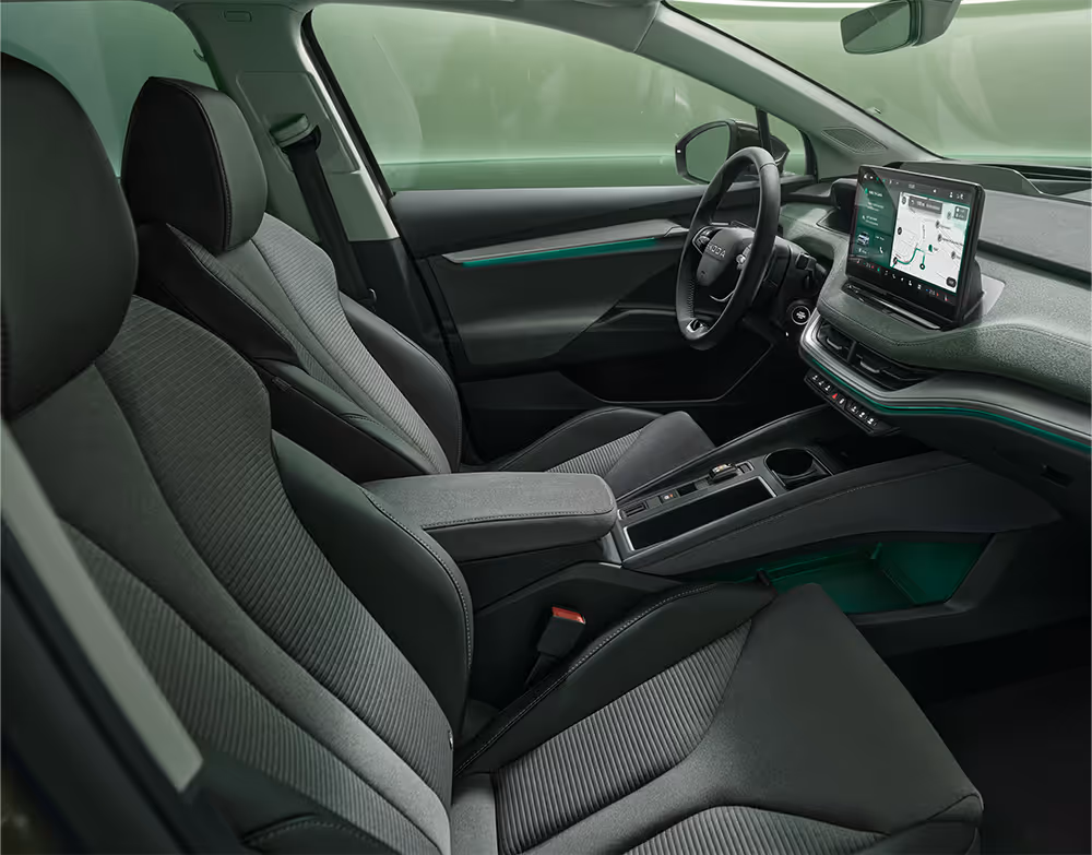 Ein Blick in den vorderen Innenraum des Škoda Enyaq mit Fokus auf den Fahrer- und Beifahrersitz. Die Sitze der Design Selection „L&K“ sind mit beigem Leder bezogen und weisen cognacfarbene Ziernähte auf. Die grüne Ambientebeleuchtung leuchtet entlang des Armaturenbretts und der Fahrertür.