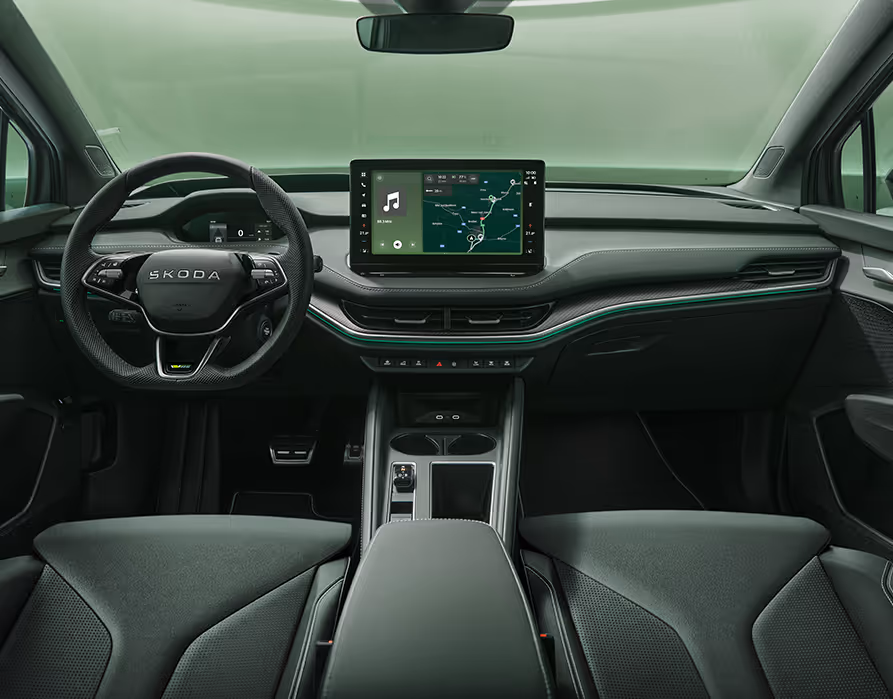 Eine symmetrische Weitwinkelaufnahme zeigt das Cockpit des Škoda Enyaq RS aus der Perspektive der Rücksitze. Das elegante Interieur der Design Selection „RS Suite“ besticht durch perforierte schwarze Lederbezüge und Dekor in Carbon-Optik sowie grüne Ambientebeleuchtung.