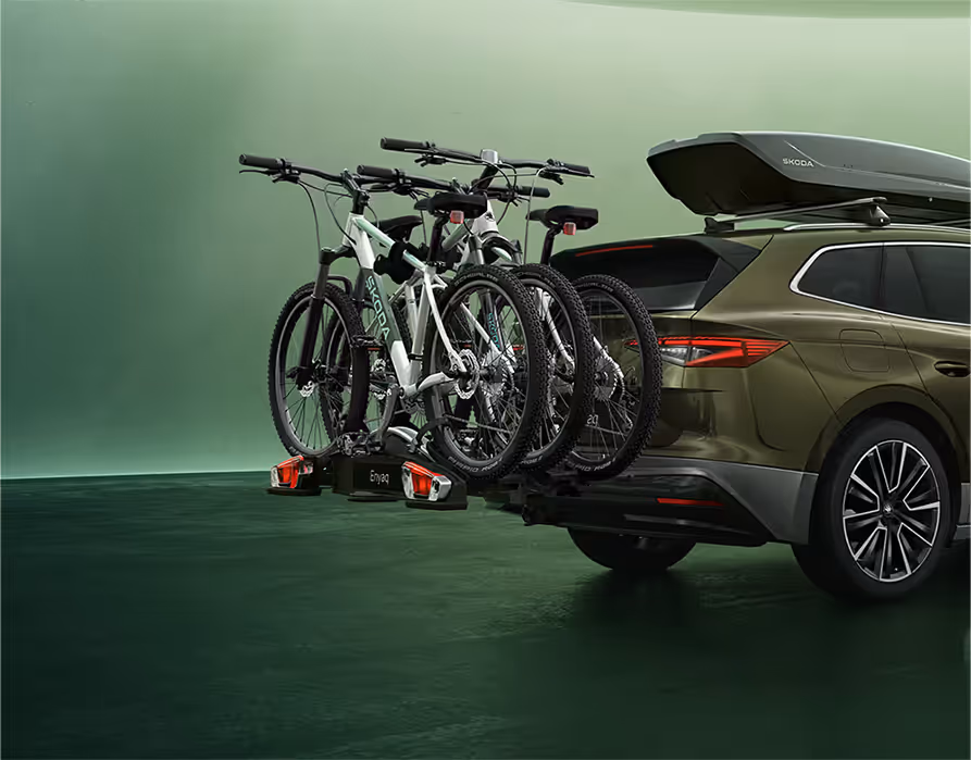 Ein Škoda Enyaq in der Lackierung Olibo-Grün Metallic wird von schräg hinten in einem grünen Studio gezeigt. Auf der Dachreling des Fahrzeugs ist eine dunkle Dachbox montiert. Zusätzlich ist am Heck ein Fahrradträger an der Anhängerkupplung befestigt, der drei Mountainbikes transportiert.