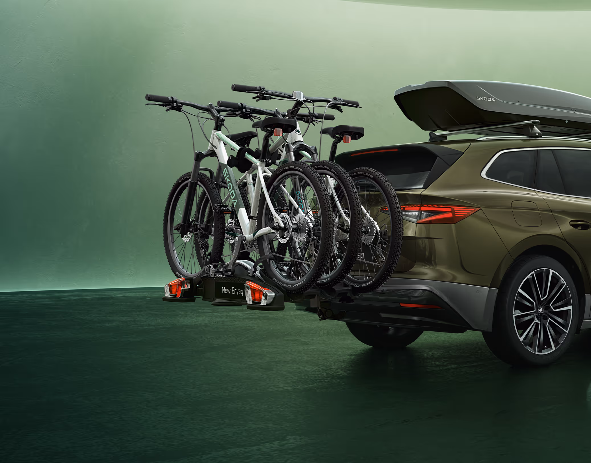 Ein Škoda Enyaq in der Lackierung Olibo-Grün Metallic wird von schräg hinten in einem grünen Studio gezeigt. Auf der Dachreling des Fahrzeugs ist eine dunkle Dachbox montiert. Zusätzlich ist am Heck ein Fahrradträger an der Anhängerkupplung befestigt, der drei Mountainbikes transportiert.