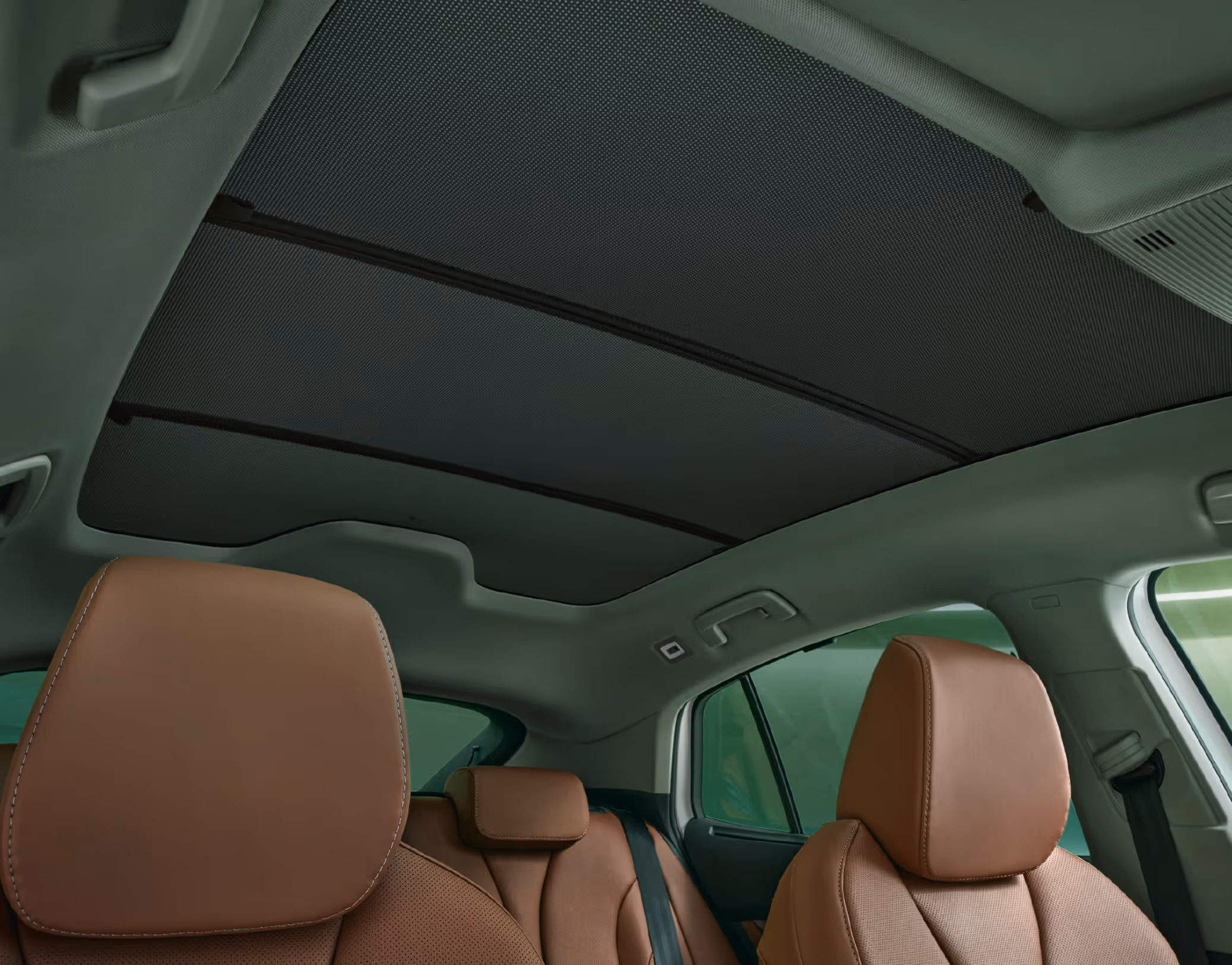 Ein Blick aus dem Innenraum eines Škoda Enyaq nach oben zum großen Panoramaglasdach. Das Dach ist mit dem elektrischen Sonnenrollo fast vollständig abgedunkelt. Im Bild sind die cognacfarbenen Ledersitze der Design Selection ecoSUITE sowie der helle Dachhimmel und die Haltegriffe zu sehen.