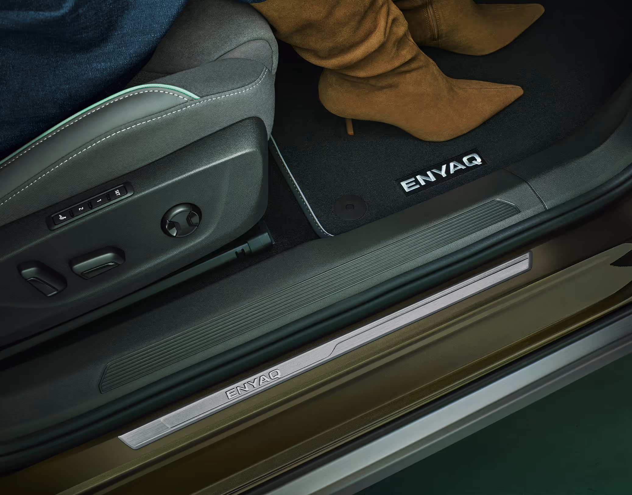 Der Beifahrerfußraum im Škoda Enyaq. Zu sehen ist der Beifahrersitz der Design Selection Lodge mit Bedienelementen für die elektrische Sitzeinstellung sowie eine Textilfußmatte mit „ENYAQ“-Schriftzug, auf der eine weibliche Person mit braunen Stiefeln ihre Füße abgestellt hat. Im geöffneten Türrahmen sind die dekorative Einstiegsleiste und ein Lautsprecher des CANTON Soundsystems erkennbar.