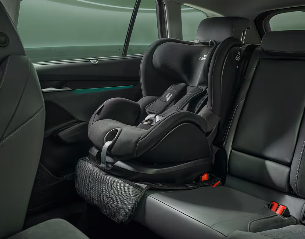 Blick auf die Rückbank eines Škoda Enyaq in der Design Selection "Loft". Auf dem rechten Sitzplatz ist ein schwarzer Baby-Kindersitz auf einer Schutzunterlage befestigt. Die Aufnahme erfasst den Bereich vom Beifahrersitz bis zur hinteren Seitentür und zeigt die Platzverhältnisse im Innenraum.