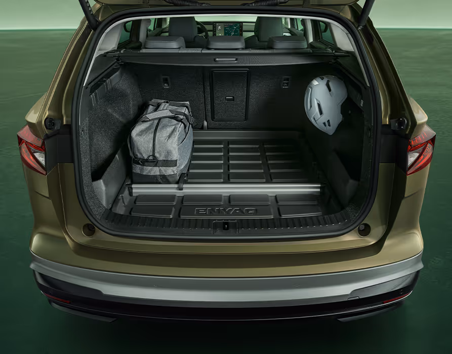 Der Blick in den geöffneten Kofferraum eines Škoda Enyaq in der Farbe Olibo-Grün Metallic. Der Kofferraumboden ist mit einer schwarzen Kunststoffwanne mit „ENYAQ“-Schriftzug ausgelegt, auf der eine graue Sporttasche. Rechts an einem Haken hängt ein grauer Fahrradhelm. Ein Aluminium-Trennelement unterteilt den vorderen und hinteren Bereich des Laderaums.