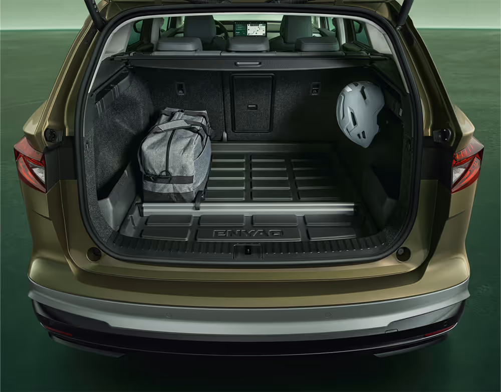 Der Blick in den geöffneten Kofferraum eines Škoda Enyaq in der Farbe Olibo-Grün Metallic. Der Kofferraumboden ist mit einer schwarzen Kunststoffwanne mit „ENYAQ“-Schriftzug ausgelegt, auf der eine graue Sporttasche. Rechts an einem Haken hängt ein grauer Fahrradhelm. Ein Aluminium-Trennelement unterteilt den vorderen und hinteren Bereich des Laderaums.