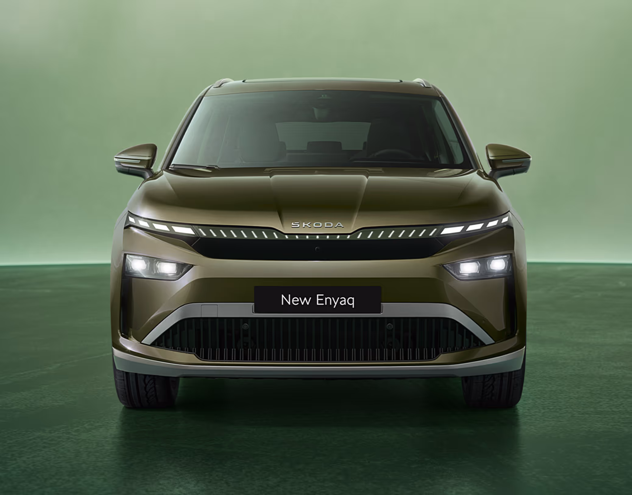 Frontalansicht des Škoda Enyaq in der Farbe Olibo-Grün Metallic vor einem grünen Studiohintergrund. Das neue Tech-Deck-Face des Fahrzeugs betont das moderne Design der Frontpartie.