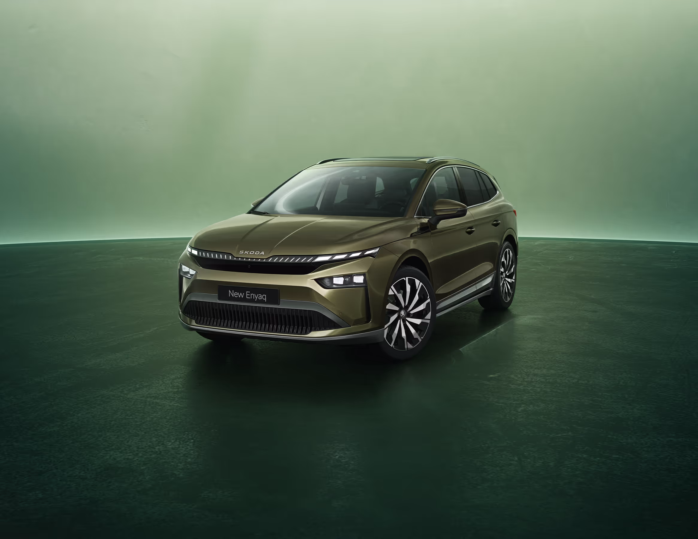 Dreiviertel-Frontansicht auf einen Škoda Enyaq in der Farbe Olibo-Grün Metallic vor einem grünen Studiohintergrund. Oben links steht geschrieben "Škoda 130 Jahre". Unten rechts ist der Schriftzug "Enyaq" platziert.