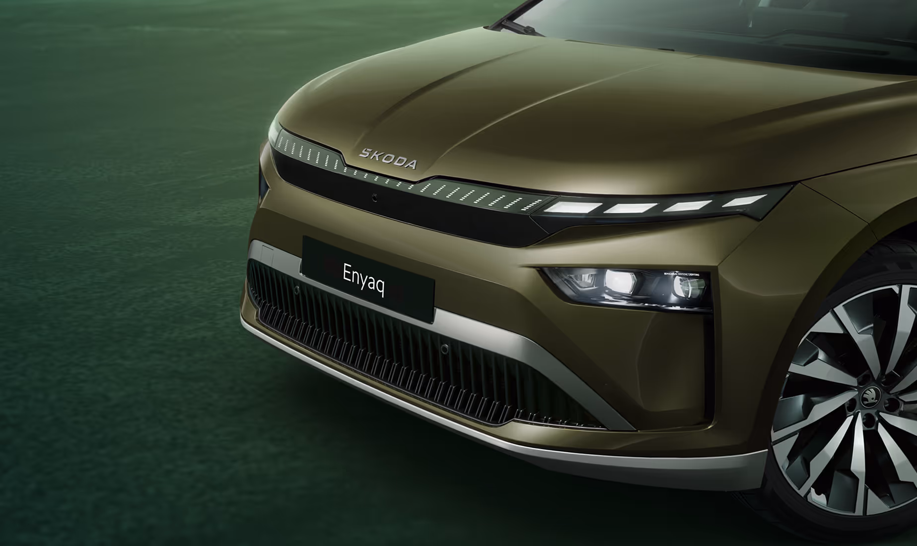 Eine Nahaufnahme der Frontpartie des Škoda Enyaq in der Farbe Olibo-Grün Metallic. Im Fokus steht der beleuchtete, markante Kühlergrill, der zusammen mit den Voll-LED-Matrix-Scheinwerfern das moderne und selbstbewusste Design unterstreicht. Unten rechts steht auf einer dunkelgrün abgesetzten Fläche der Schriftzug „Sicherheit und Assistenten“.