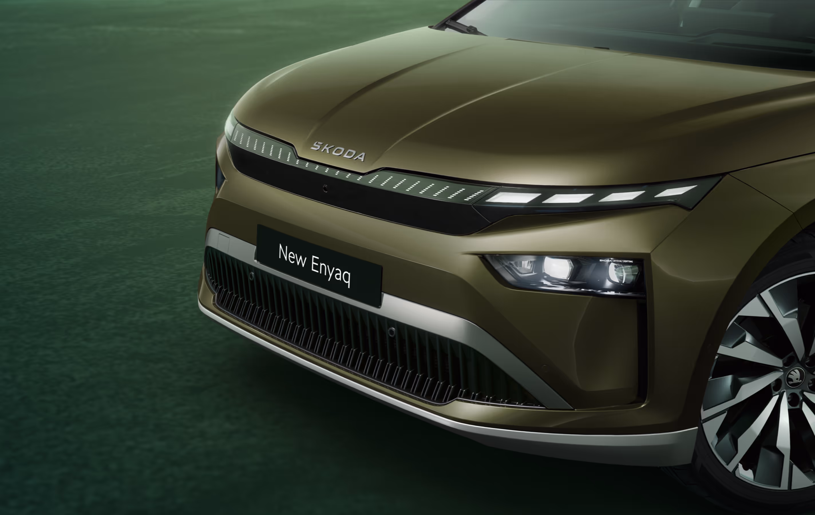 Eine Nahaufnahme der Frontpartie des Škoda Enyaq in der Farbe Olibo-Grün Metallic. Im Fokus steht der beleuchtete, markante Kühlergrill, der zusammen mit den Voll-LED-Matrix-Scheinwerfern das moderne und selbstbewusste Design unterstreicht. Unten rechts steht auf einer dunkelgrün abgesetzten Fläche der Schriftzug „Sicherheit und Assistenten“.