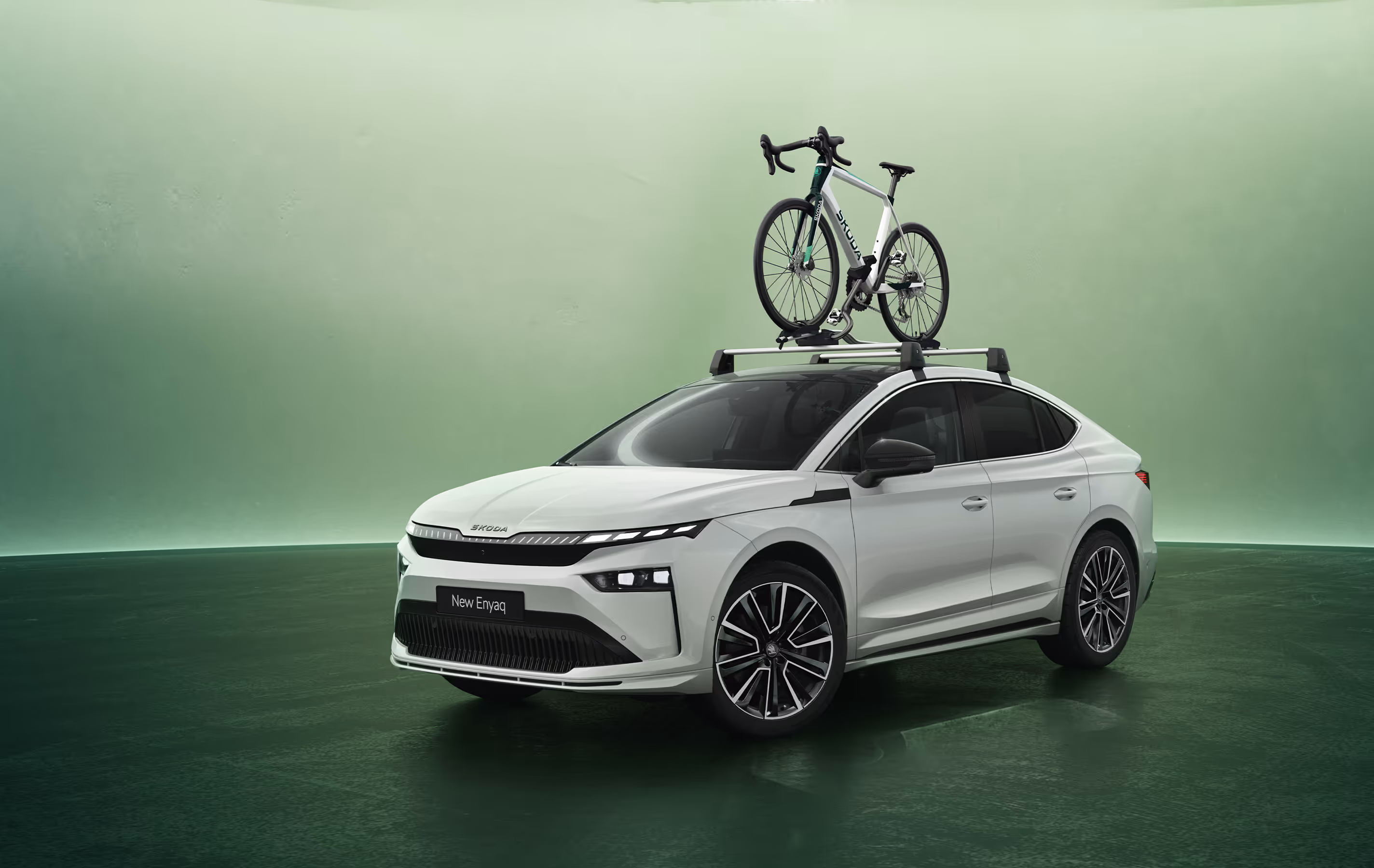 Ein weißer Škoda Enyaq Coupé mit beleuchtetem Crystal Face Kühlergrill steht schräg von vorn in einem Studio mit grünem Hintergrund. Auf dem Fahrzeugdach ist ein Dachträger montiert, auf dem ein Rennrad befestigt ist. Unten rechts im Bild steht auf einer dunkelgrün abgesetzten Fläche der Schriftzug "Škoda Original Zubehör".
