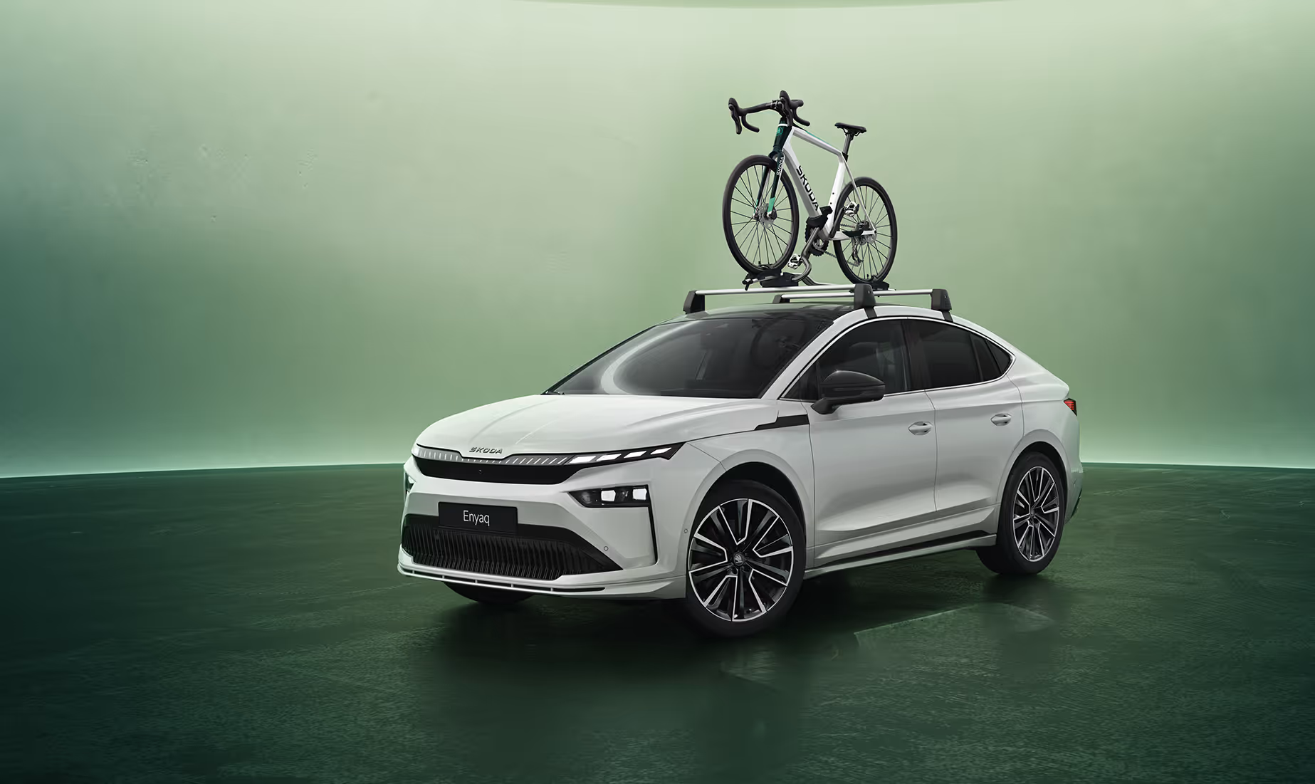 Ein weißer Škoda Enyaq Coupé mit beleuchtetem Crystal Face Kühlergrill steht schräg von vorn in einem Studio mit grünem Hintergrund. Auf dem Fahrzeugdach ist ein Dachträger montiert, auf dem ein Rennrad befestigt ist. Unten rechts im Bild steht auf einer dunkelgrün abgesetzten Fläche der Schriftzug "Škoda Original Zubehör".