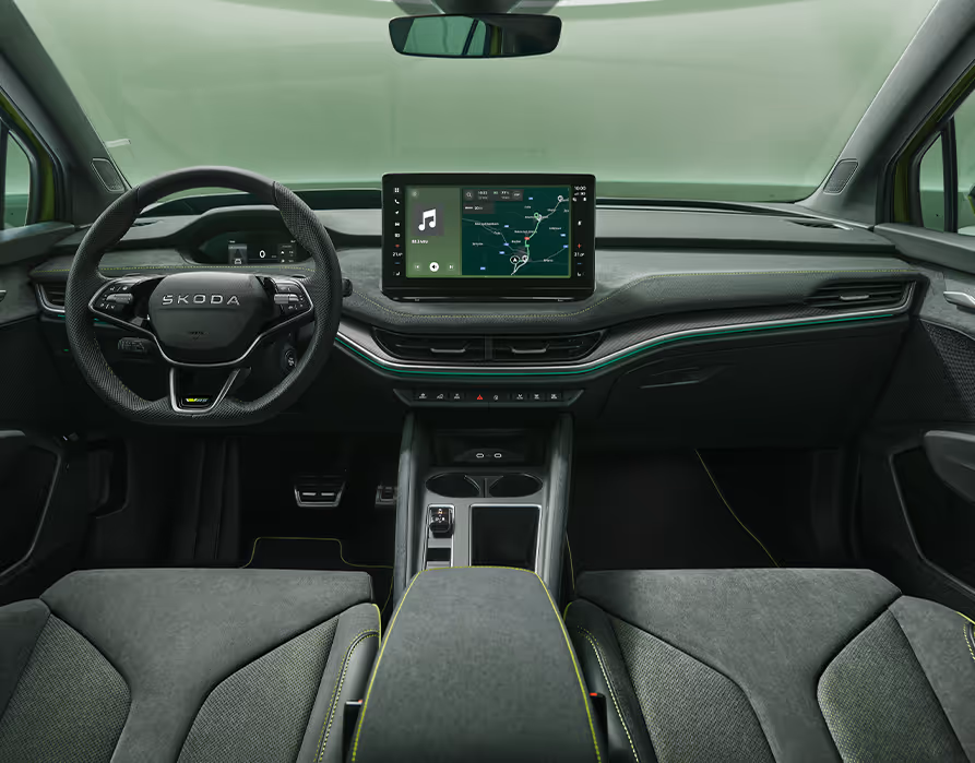 Eine symmetrische Weitwinkelaufnahme zeigt das Cockpit des Škoda Enyaq RS aus der Perspektive der Rücksitze. Das elegante Interieur der Design Selection „RS Lounge“ besticht durch perforierte schwarze Lederbezüge und Dekor in Carbon-Optik sowie grüne Ambientebeleuchtung.
