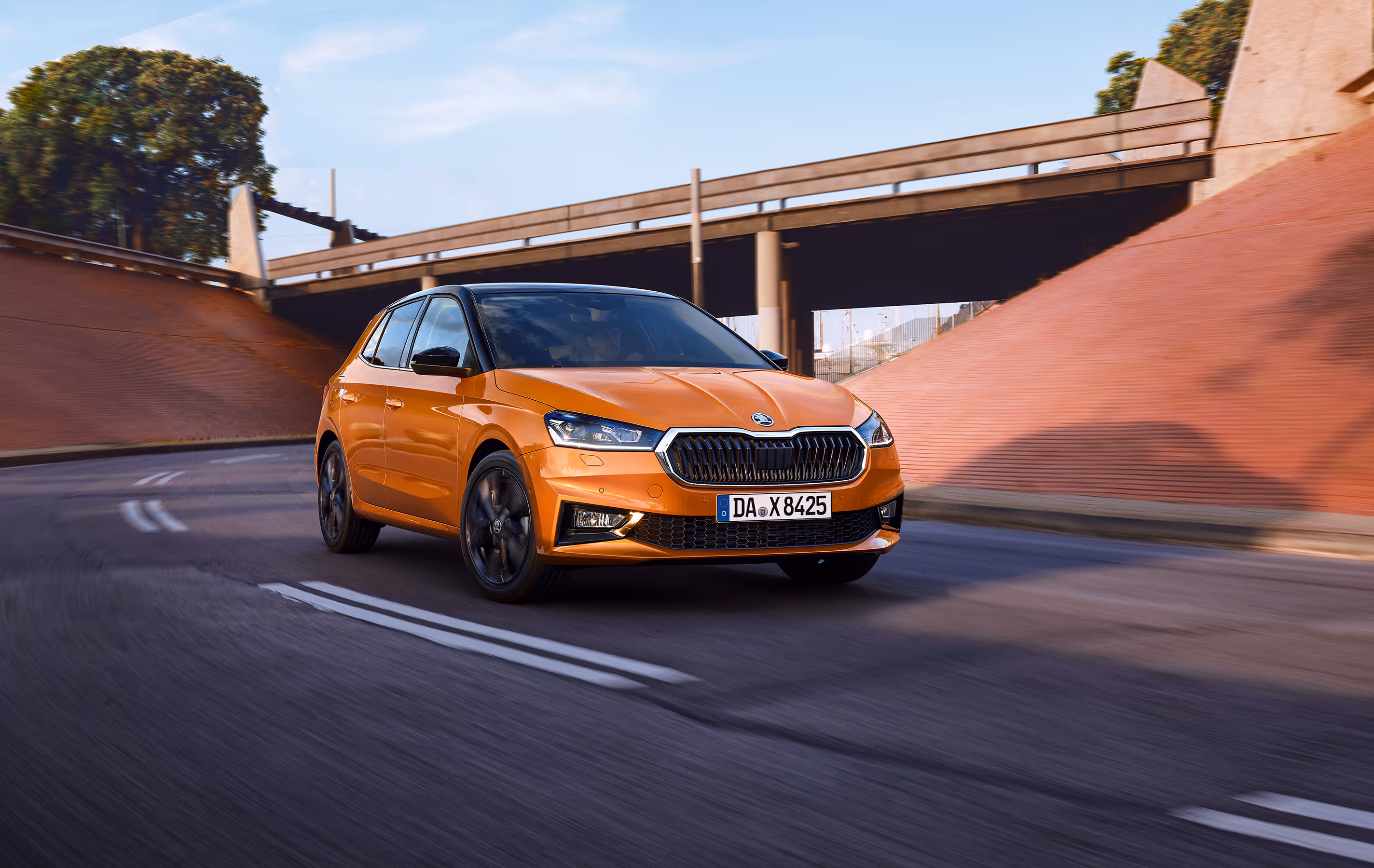 Schräge Frontalansicht eines fahrenden Škoda Fabia in der Lackierung Phoenix-Orange Metallic. Das Fahrzeug fährt dynamisch auf einer asphaltierten Straße unter einer Brückenunterführung hindurch. Ausgestattet ist es mit dem „Color Concept Schwarz“, erkennbar am kontrastierenden schwarzen Dach sowie den schwarzen Außenspiegelkappen.