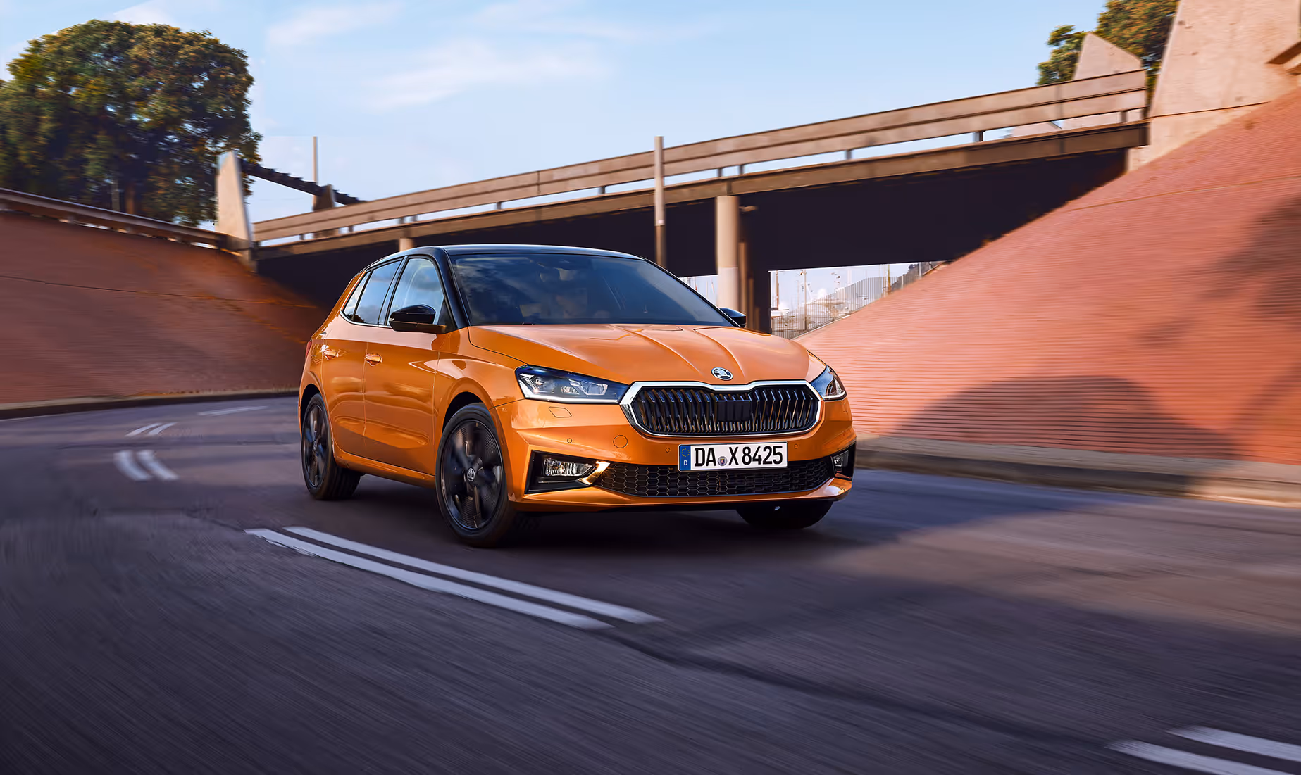 Schräge Frontalansicht eines fahrenden Škoda Fabia in der Lackierung Phoenix-Orange Metallic. Das Fahrzeug fährt dynamisch auf einer asphaltierten Straße unter einer Brückenunterführung hindurch. Ausgestattet ist es mit dem „Color Concept Schwarz“, erkennbar am kontrastierenden schwarzen Dach sowie den schwarzen Außenspiegelkappen.