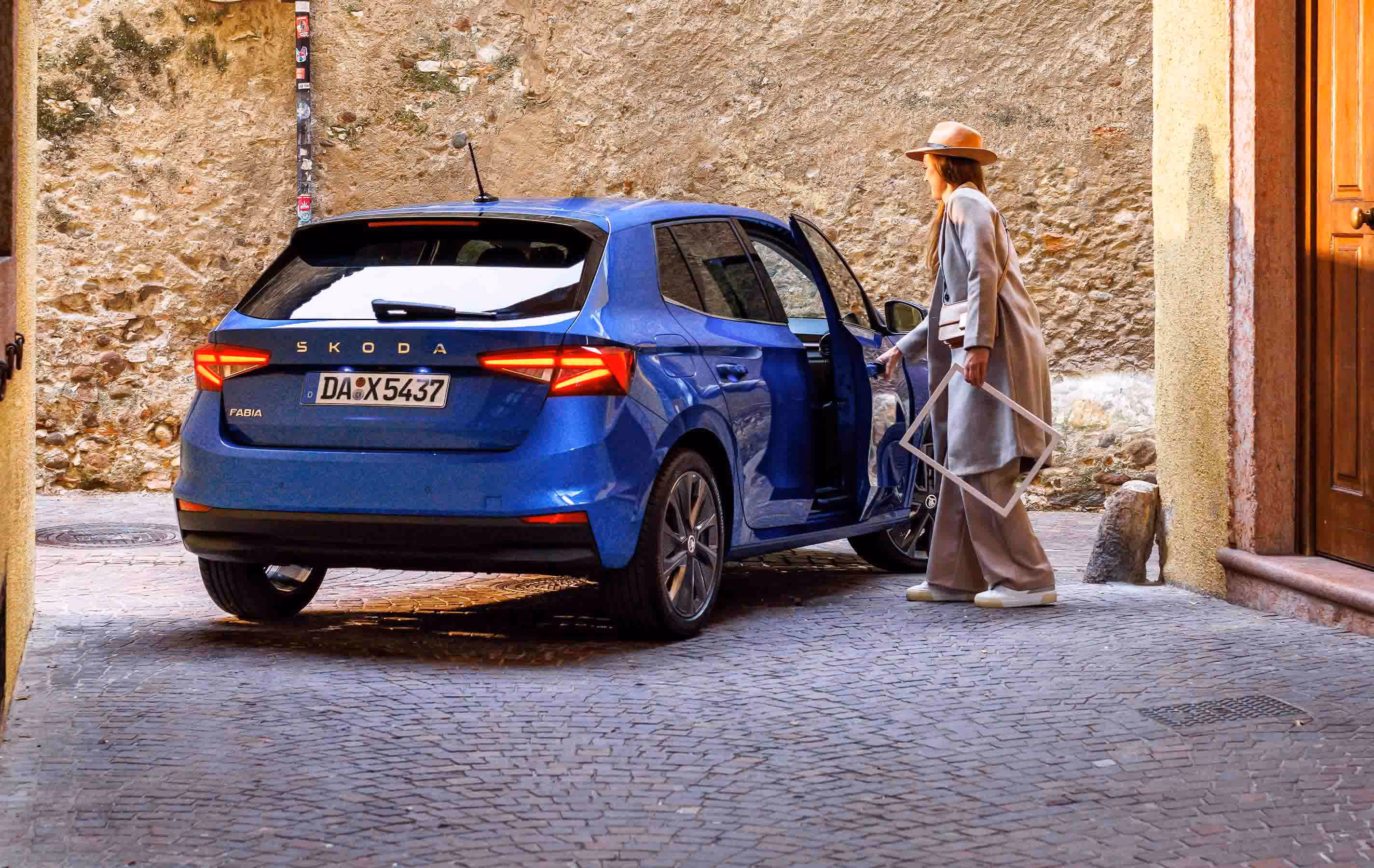 Heckansicht eines stehenden Škoda Fabia in der Lackierung Race-Blau Metallic in einer engen Gasse mit Kopfsteinpflaster und historischen Steinmauern. Eine Frau, bekleidet mit einem beigen Mantel und Hut, öffnet gerade die Beifahrertür und hält dabei einen leeren, weißen Bilderrahmen in der Hand. Die markanten LED-Heckleuchten des Fahrzeugs sind aktiviert.