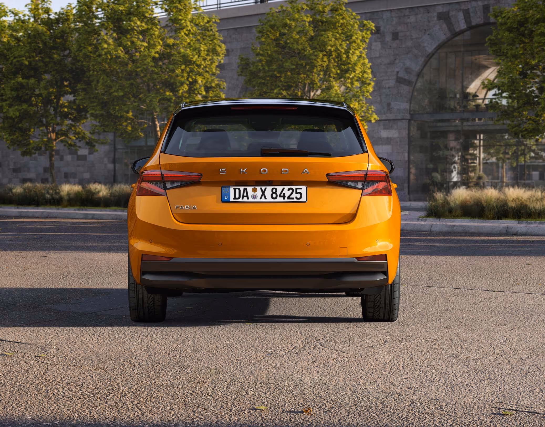 Heckansicht eines Škoda Fabia in der Lackierung Phoenix-Orange Metallic, der vor einer städtischen Kulisse mit Bäumen und einer Eisenbahnbrücke aus Naturstein geparkt ist. Das Fahrzeug ist mit dem Color Concept ausgestattet, was am schwarz lackierten Dach sowie den schwarzen Außenspiegelkappen erkennbar ist. Der zentrale Škoda-Schriftzug auf der Heckklappe und die markanten Heckleuchten runden das Design ab.