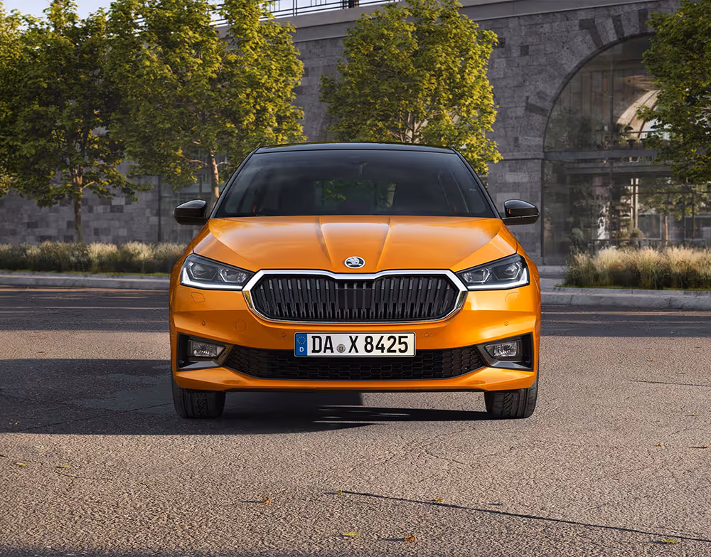 Frontansicht eines Škoda Fabia in der Lackierung Phoenix-Orange Metallic, der vor einer städtischen Kulisse mit Bäumen und einer Eisenbahnbrücke aus Naturstein geparkt ist. Das Fahrzeug ist mit dem Color Concept ausgestattet, was am schwarz lackierten Dach sowie den schwarzen Außenspiegelkappen erkennbar ist, und zeigt seine markante Frontpartie mit LED-Scheinwerfern.