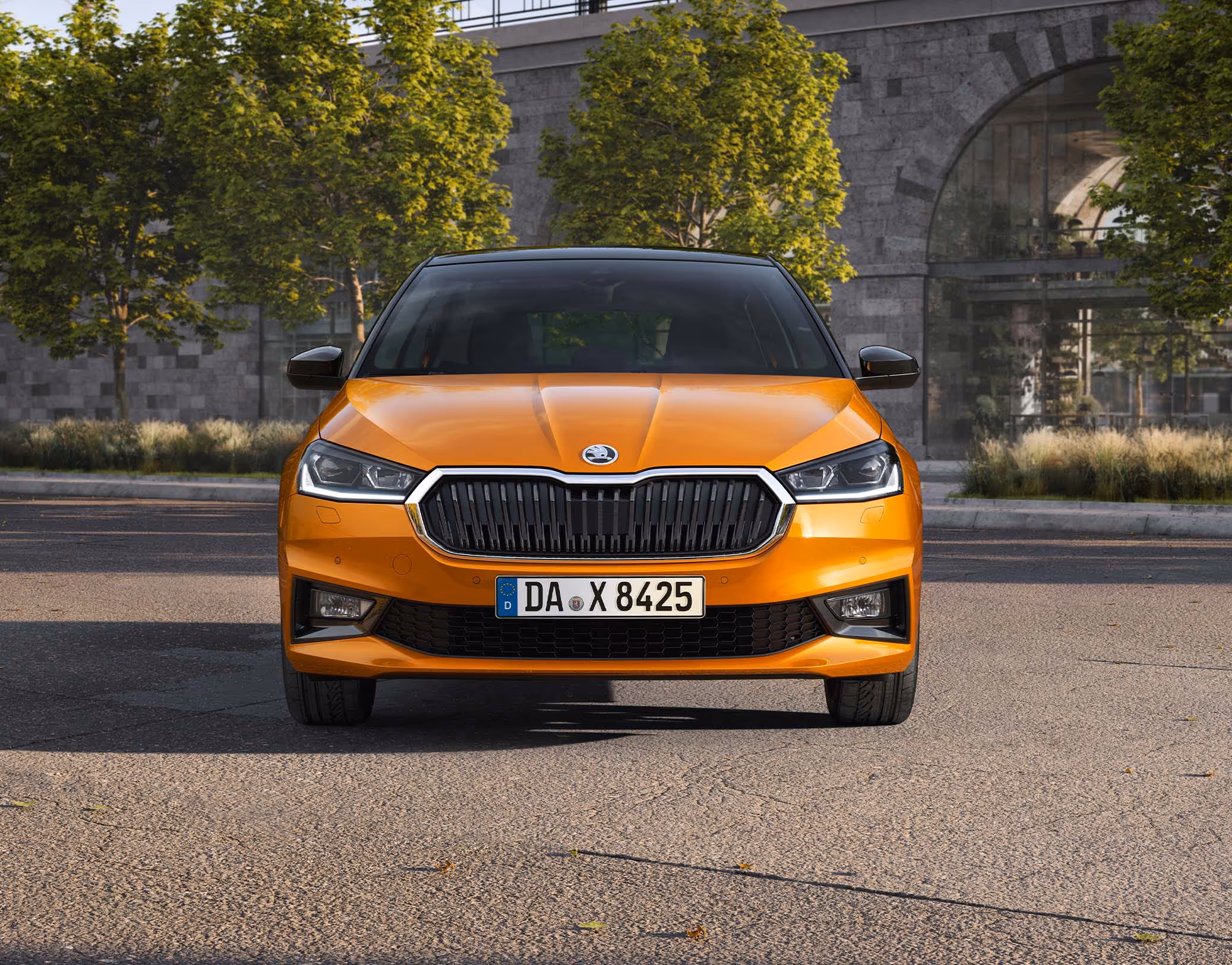 Frontansicht eines Škoda Fabia in der Lackierung Phoenix-Orange Metallic, der vor einer städtischen Kulisse mit Bäumen und einer Eisenbahnbrücke aus Naturstein geparkt ist. Das Fahrzeug ist mit dem Color Concept ausgestattet, was am schwarz lackierten Dach sowie den schwarzen Außenspiegelkappen erkennbar ist, und zeigt seine markante Frontpartie mit LED-Scheinwerfern.