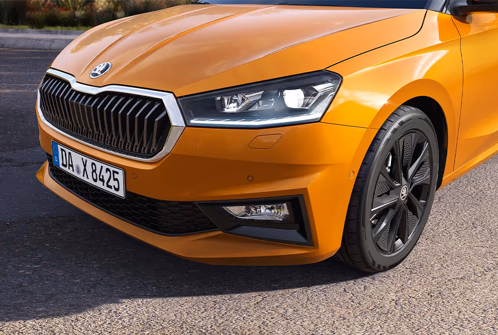 Detailansicht der Frontpartie eines Škoda Fabia in der Lackierung Phoenix-Orange Metallic, geparkt auf asphaltiertem Untergrund. Im Fokus stehen der scharf gezeichnete LED-Scheinwerfer, der Kühlergrill mit Chromrahmen sowie die schwarze Leichtmetallfelge mit Aero-Blenden. Die Aufnahme hebt zudem die integrierten Nebelscheinwerfer und die markanten Linien der Motorhaube hervor.