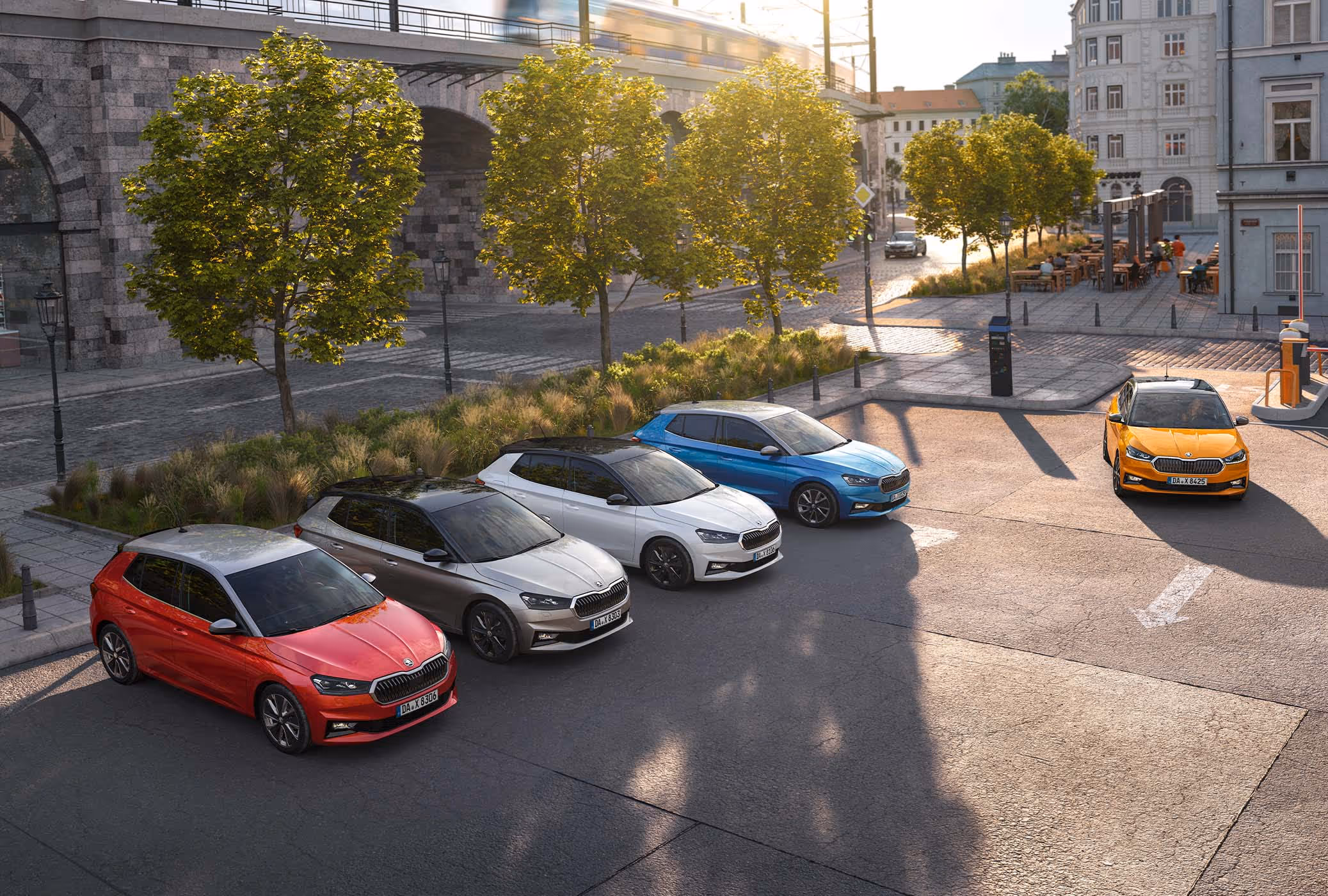 Vogelperspektive auf fünf Škoda Fabia in unterschiedlichen Lackierungen, die fächerförmig auf einem städtischen Platz stehen. Alle Fahrzeuge sind mit dem Color Concept ausgestattet, was an den kontrastierenden Dachfarben in Schwarz oder Grau erkennbar ist. Im sonnendurchfluteten Hintergrund befindet sich eine Eisenbahnbrücke aus Naturstein mit umgebenden Bäumen.