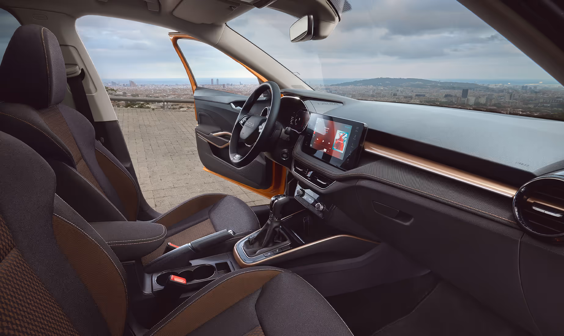 Blick in den Innenraum eines Škoda Fabia, der durch Stoff-Sitzbezüge mit Kontrastnähten und eine markante Dekorleiste in Copper veredelt ist. Das moderne Cockpit umfasst ein freistehendes Infotainment-Display sowie ein digitales Kombiinstrument hinter dem Zweispeichen-Multifunktionslenkrad. Durch die geöffnete Fahrertür und die Windschutzscheibe bietet sich ein Ausblick auf eine weitläufige Stadtkulisse.