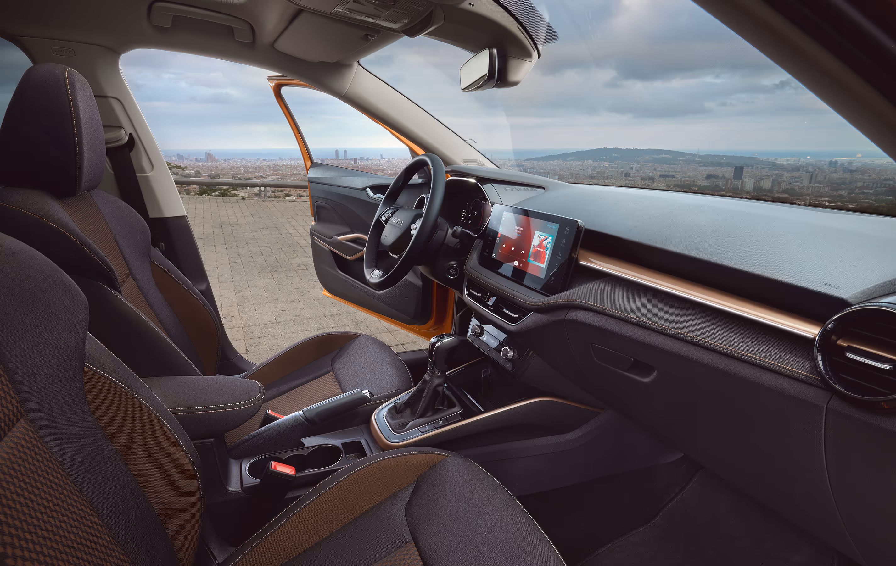 Blick in den Innenraum eines Škoda Fabia, der durch Stoff-Sitzbezüge mit Kontrastnähten und eine markante Dekorleiste in Copper veredelt ist. Das moderne Cockpit umfasst ein freistehendes Infotainment-Display sowie ein digitales Kombiinstrument hinter dem Zweispeichen-Multifunktionslenkrad. Durch die geöffnete Fahrertür und die Windschutzscheibe bietet sich ein Ausblick auf eine weitläufige Stadtkulisse.
