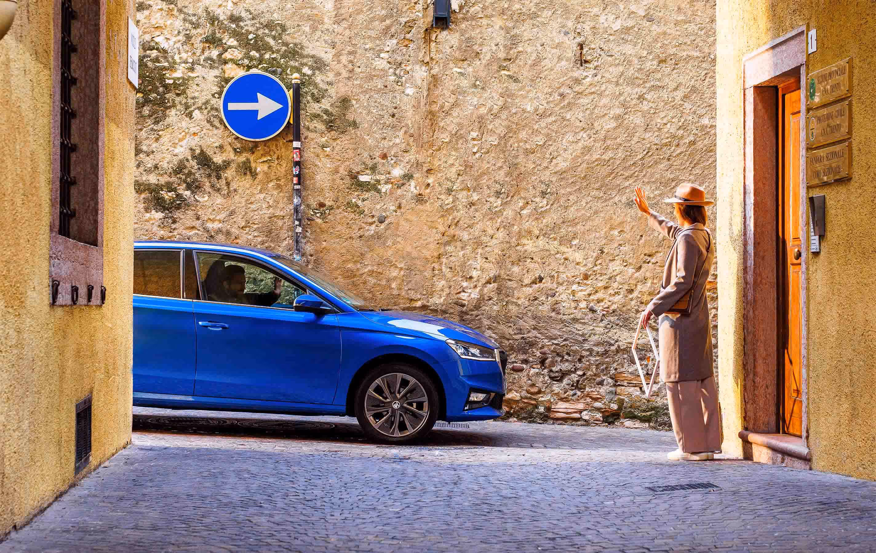 Seitenansicht eines Škoda Fabia in der Lackierung Race-Blau Metallic, der durch eine enge Gasse mit historischen Steinmauern fährt. Eine Frau mit Hut steht am Straßenrand und winkt dem Fahrer zu, der den Gruß erwidert. Ein blaues Verkehrsschild an der Mauer weist die Fahrtrichtung in der malerischen Altstadtgasse.