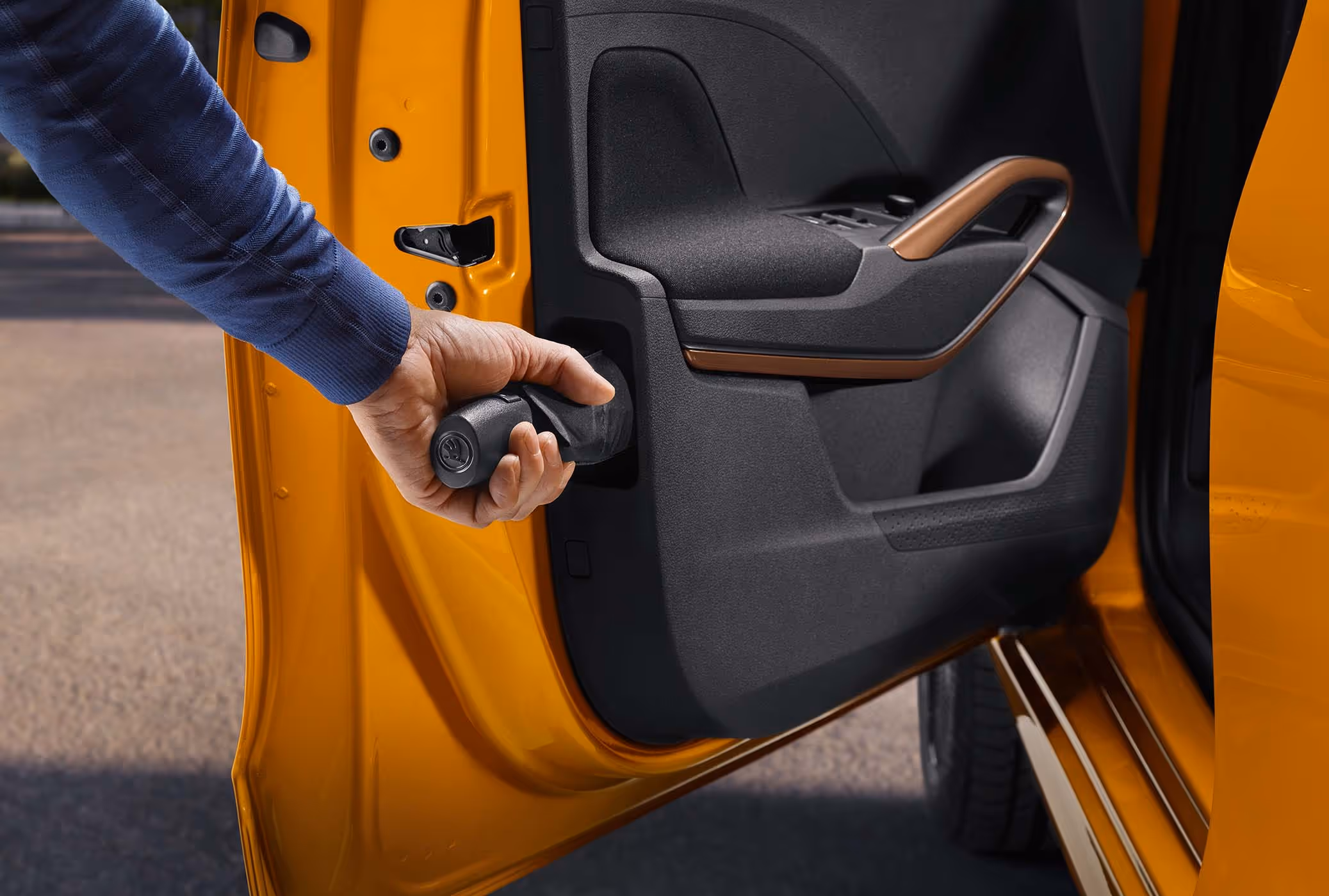 Detailansicht der geöffneten Tür eines Škoda Fabia in Phoenix-Orange Metallic, bei der eine Hand einen Regenschirm aus dem integrierten Ablagefach in der Türverkleidung zieht. Dieses praktische „Simply Clever“-Feature ermöglicht das griffbereite Verstauen eines Regenschirms direkt im Fahrzeug. Akzente setzt zudem der Türzuziehgriff in der Farbe Copper, der sich harmonisch in die dunkle Innenverkleidung einfügt.