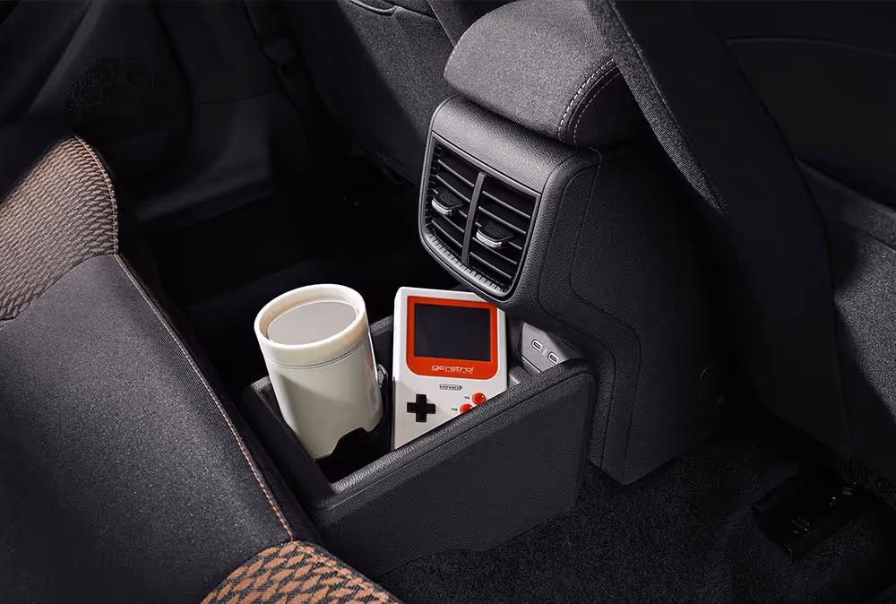 Detailansicht der hinteren Mittelkonsole eines Škoda Fabia, die eine flexible Ablagebox auf dem Mitteltunnel zeigt, in der ein Becher und eine Spielkonsole Platz finden. Darüber sind zwei USB-C-Ladeanschlüsse sowie verstellbare Lüftungsdüsen für die Fondpassagiere angeordnet. Angeschnitten ist zudem der Rücksitz mit den charakteristischen Stoffbezügen und Kontrastnähten in Copper.