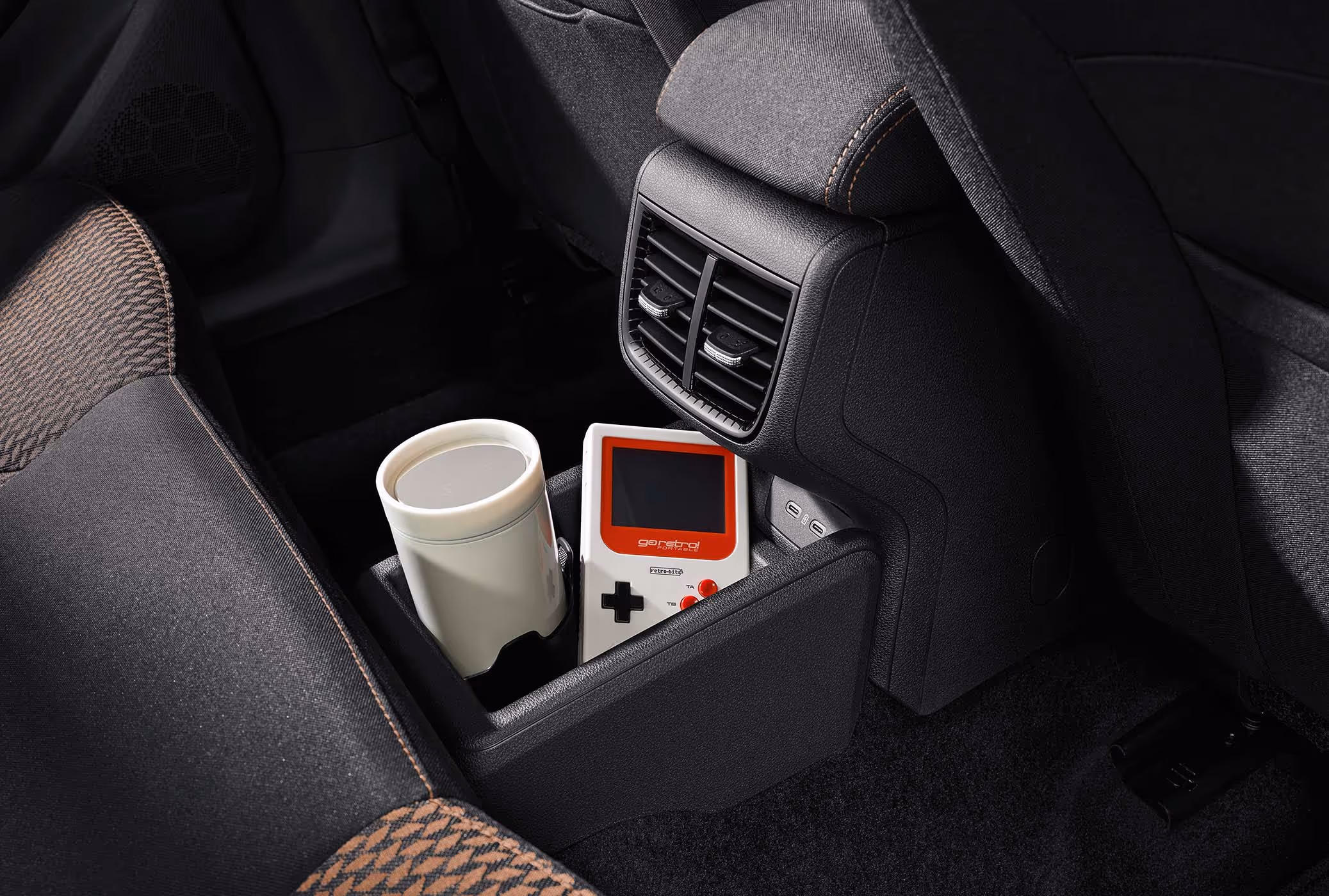 Detailansicht der hinteren Mittelkonsole eines Škoda Fabia, die eine flexible Ablagebox auf dem Mitteltunnel zeigt, in der ein Becher und eine Spielkonsole Platz finden. Darüber sind zwei USB-C-Ladeanschlüsse sowie verstellbare Lüftungsdüsen für die Fondpassagiere angeordnet. Angeschnitten ist zudem der Rücksitz mit den charakteristischen Stoffbezügen und Kontrastnähten in Copper.