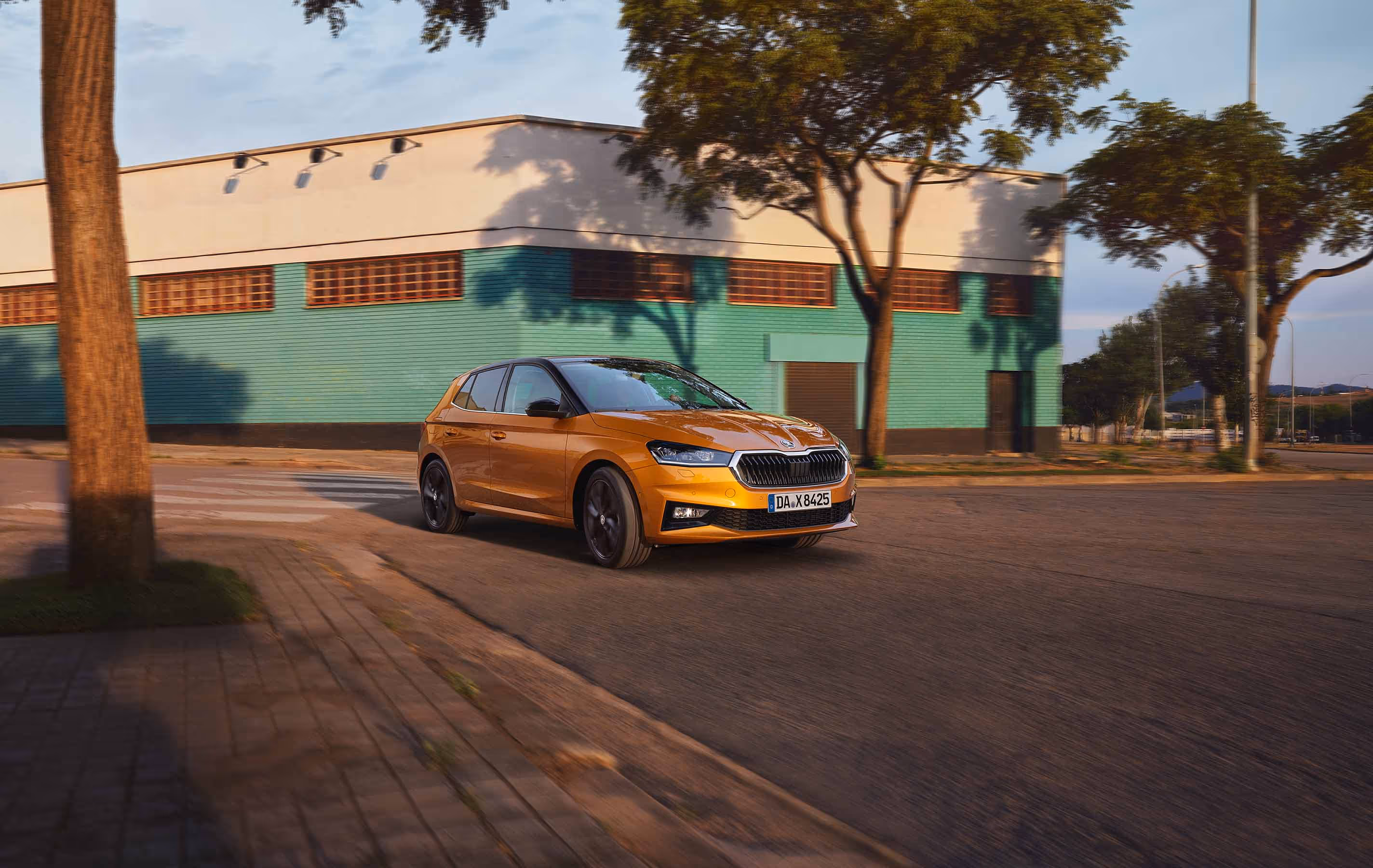 Ein Škoda Fabia in der Lackierung Phoenix-Orange Metallic fährt dynamisch auf einer Straße vor einem Gebäude mit türkisfarbener Fassade. Das Fahrzeug ist mit einem kontrastierenden schwarzen Dach sowie schwarzen Leichtmetallfelgen ausgestattet, die das sportliche Design unterstreichen. Die städtische Umgebung im Hintergrund wird durch Bäume und das Spiel von Licht und Schatten ergänzt.