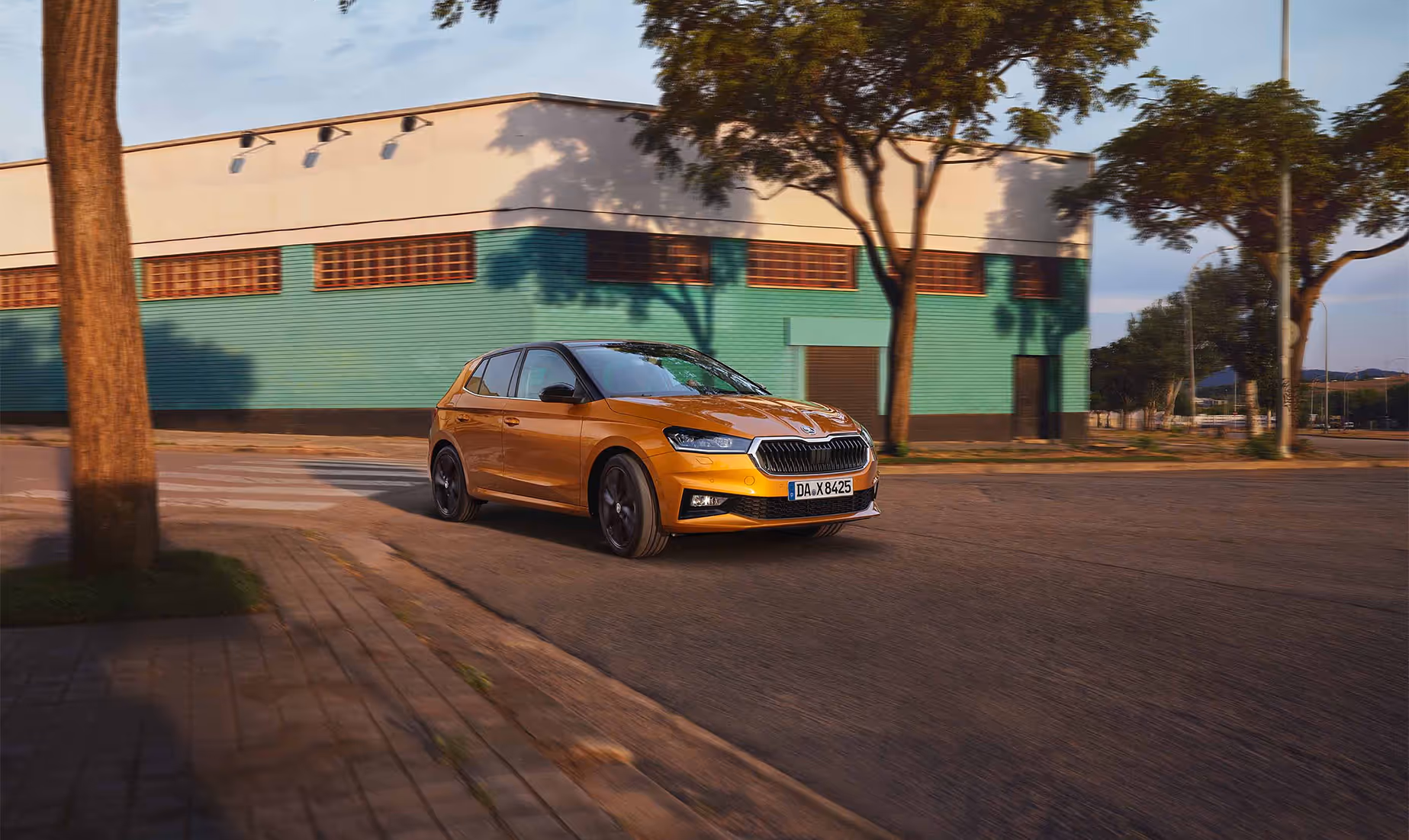 Ein Škoda Fabia in der Lackierung Phoenix-Orange Metallic fährt dynamisch auf einer Straße vor einem Gebäude mit türkisfarbener Fassade. Das Fahrzeug ist mit einem kontrastierenden schwarzen Dach sowie schwarzen Leichtmetallfelgen ausgestattet, die das sportliche Design unterstreichen. Die städtische Umgebung im Hintergrund wird durch Bäume und das Spiel von Licht und Schatten ergänzt.
