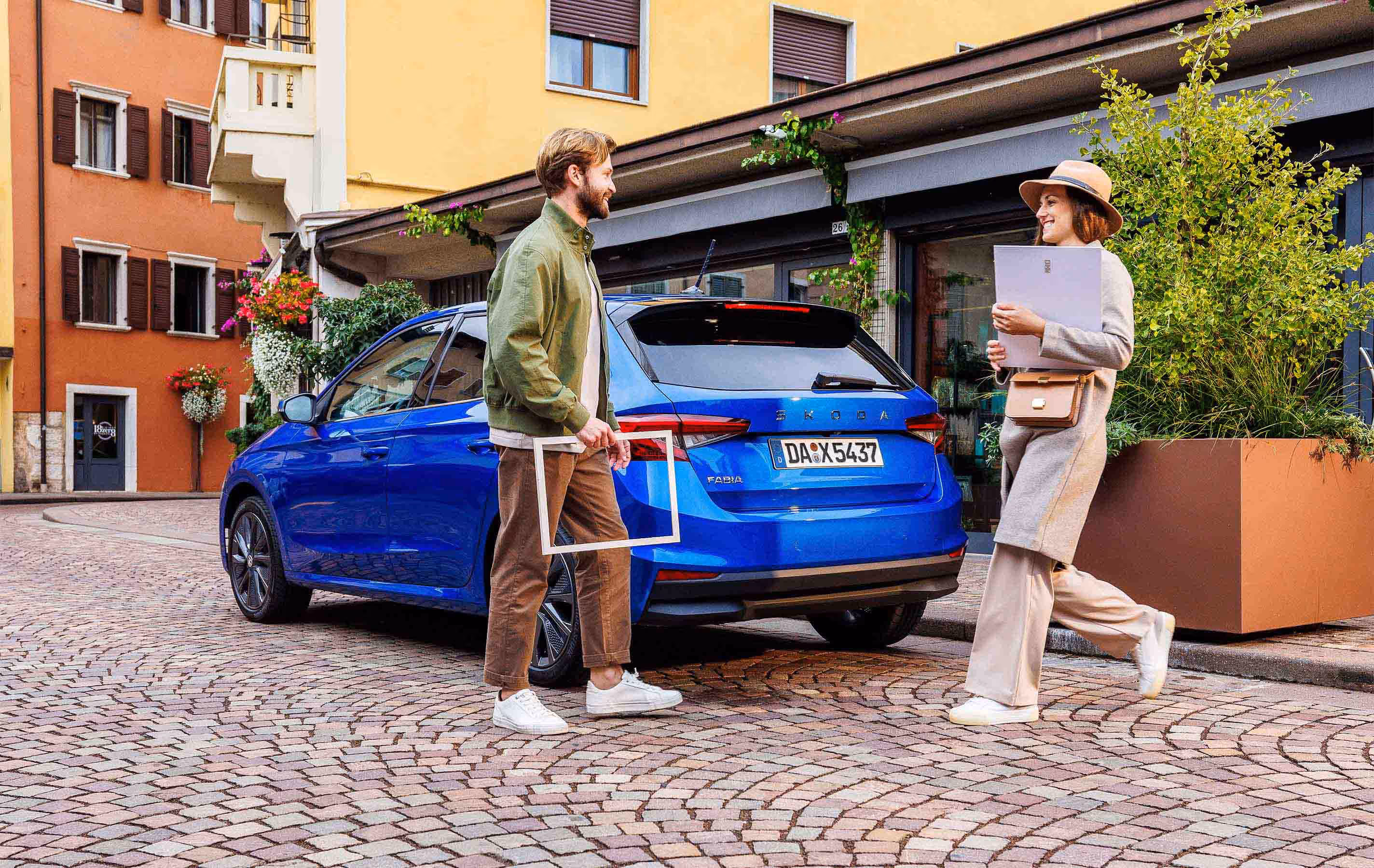 Ein Mann mit einem Holzrahmen und eine Frau mit einer Mappe begegnen sich hinter dem Heck eines geparkten Škoda Fabia in der Lackierung Race-Blau Metallic. Das Fahrzeug steht auf Kopfsteinpflaster in einer engen Gasse, umgeben von historischen Hausfassaden und Pflanzkübeln. Der Fokus liegt auf der Rückansicht des Kleinwagens mit dem markanten Škoda Schriftzug auf der Heckklappe.