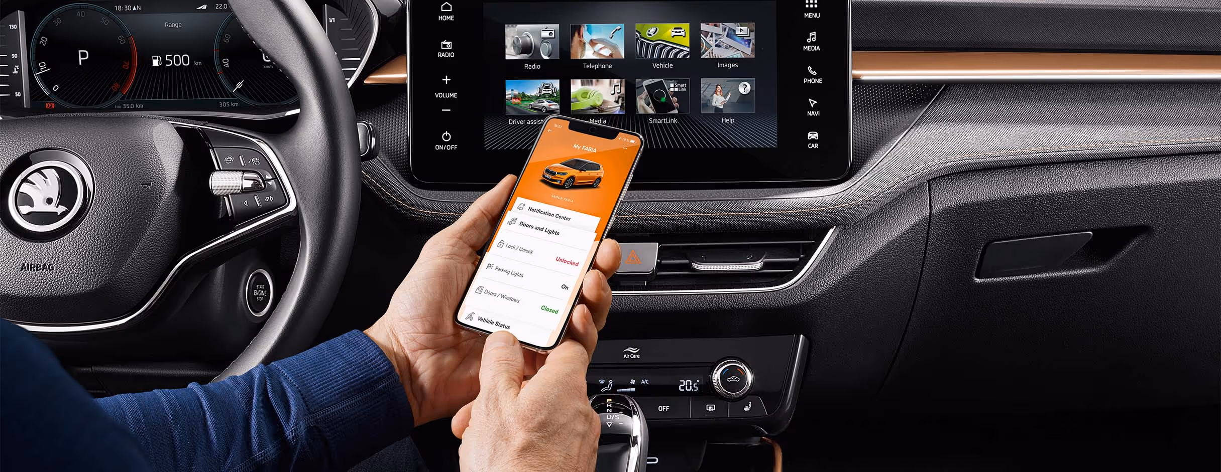 Ein Nutzer hält ein Smartphone in der Hand, auf dem die MyŠkoda App detaillierte Informationen zum Fahrzeugstatus wie Verriegelung und Beleuchtung anzeigt. Dahinter präsentiert das große Infotainment-Display das Hauptmenü mit Kacheln für Medien, Telefonie und SmartLink. Abgerundet wird das Cockpit durch das Multifunktionslenkrad und die darunterliegende Bedieneinheit der Climatronic.