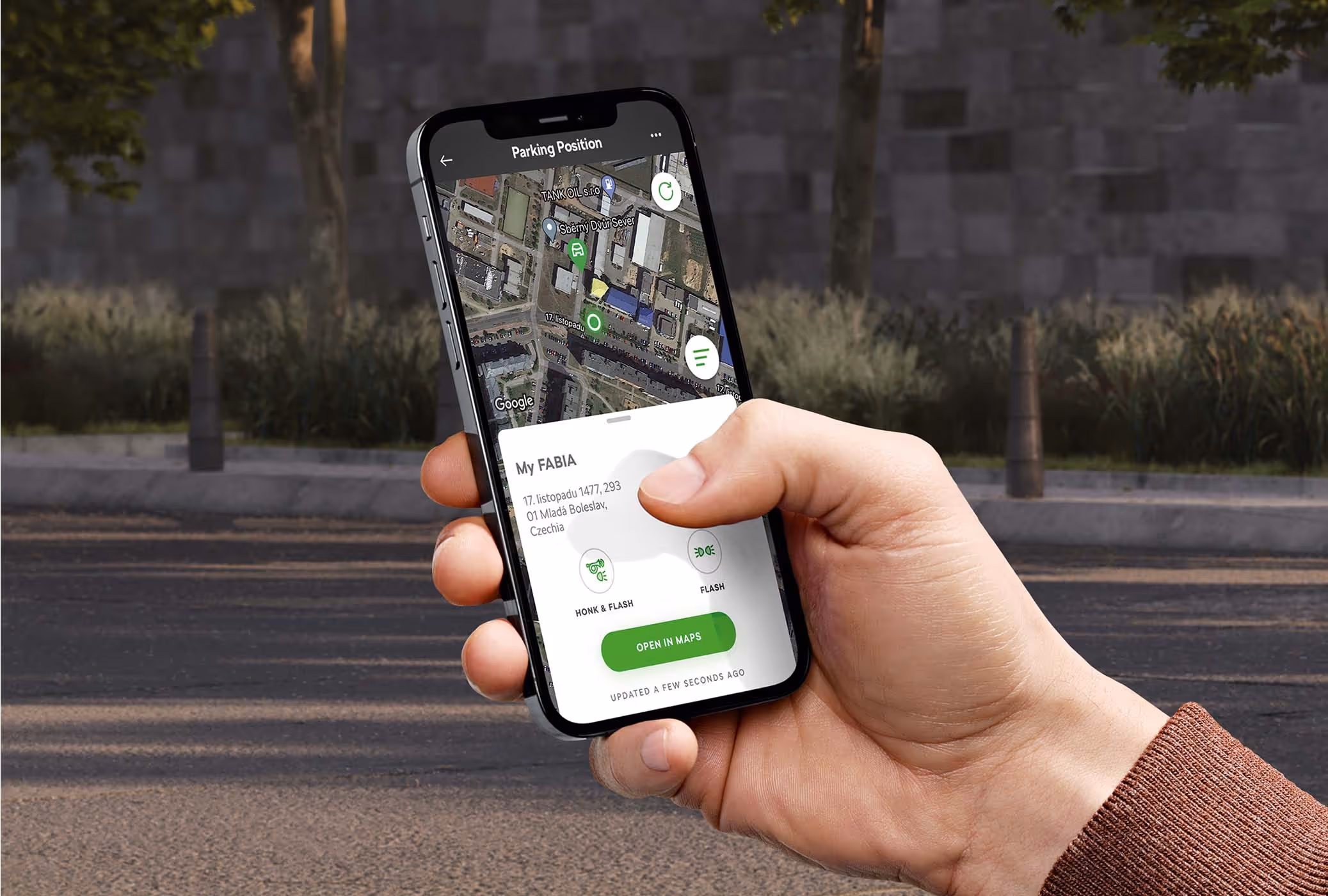 Auf dem Display eines Smartphones wird die Funktion "Parkposition" innerhalb der MyŠkoda App dargestellt. Eine Karte markiert den genauen Standort des Fahrzeugs und bietet über Schaltflächen wie "Hupen & Blinken" Unterstützung beim Auffinden. Gehalten wird das Mobiltelefon vor einer unscharfen Straßenkulisse.