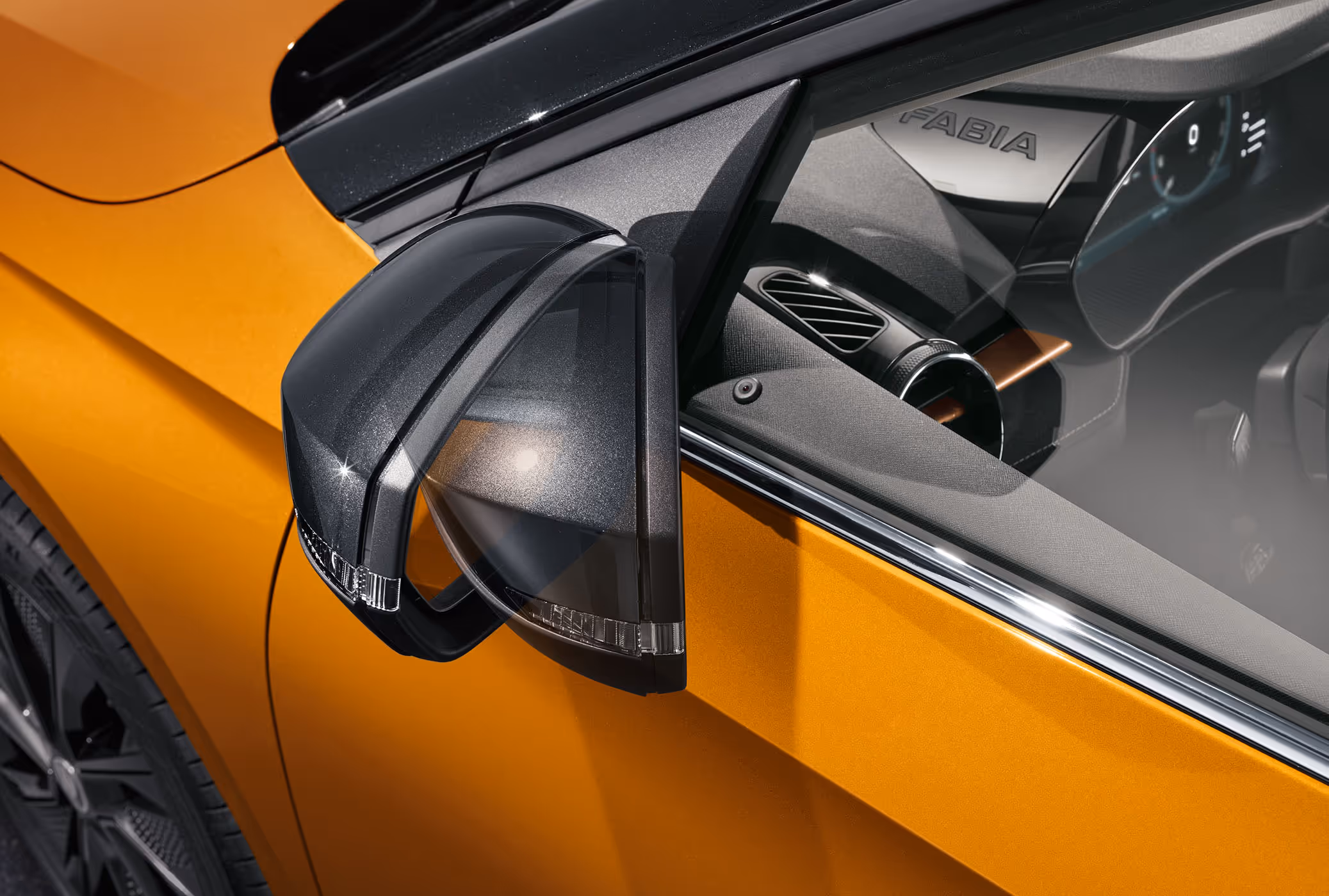 Der elektrisch anklappbare Außenspiegel des Škoda Fabia in Phoenix-Orange Metallic wird hier grafisch während des Einklappvorgangs dargestellt. Die schwarze Spiegelkappe bildet dabei einen Kontrast zur leuchtenden Lackierung und unterstreicht das dynamische Design. Durch die Seitenscheibe ist zudem ein Teil des Cockpits mit dem markanten Fabia-Schriftzug auf der Instrumentenhutze erkennbar.