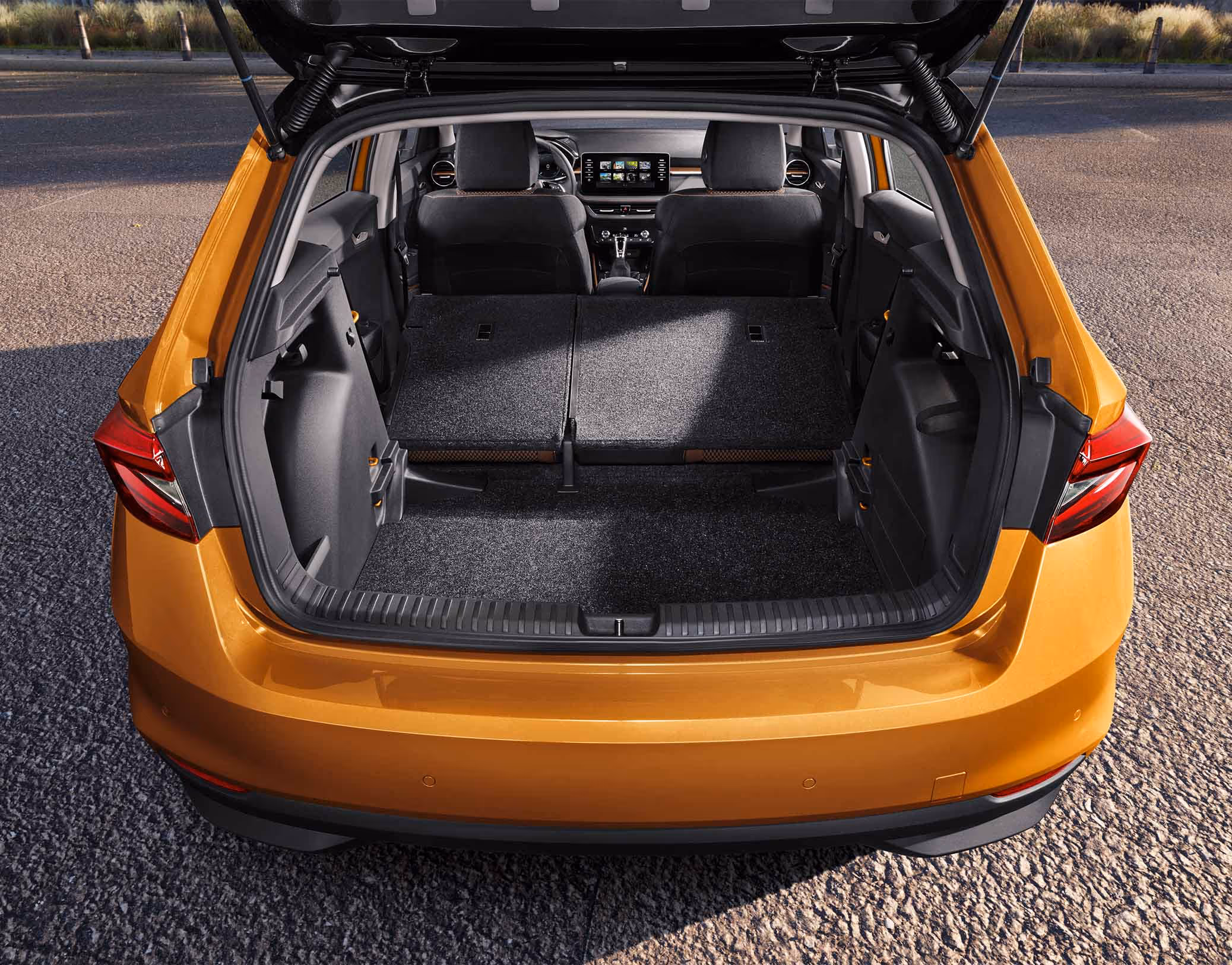Eine Heckansicht zeigt den geöffneten Kofferraum eines Škoda Fabia in der Lackierung Phoenix-Orange Metallic. Die geteilte Rücksitzbank ist vollständig umgeklappt, wodurch eine vergrößerte Ladefläche für den Transport von Gegenständen entsteht. Im vorderen Bereich sind die aufrechten Vordersitze sowie das Armaturenbrett mit dem zentralen Infotainment-Display zu sehen.