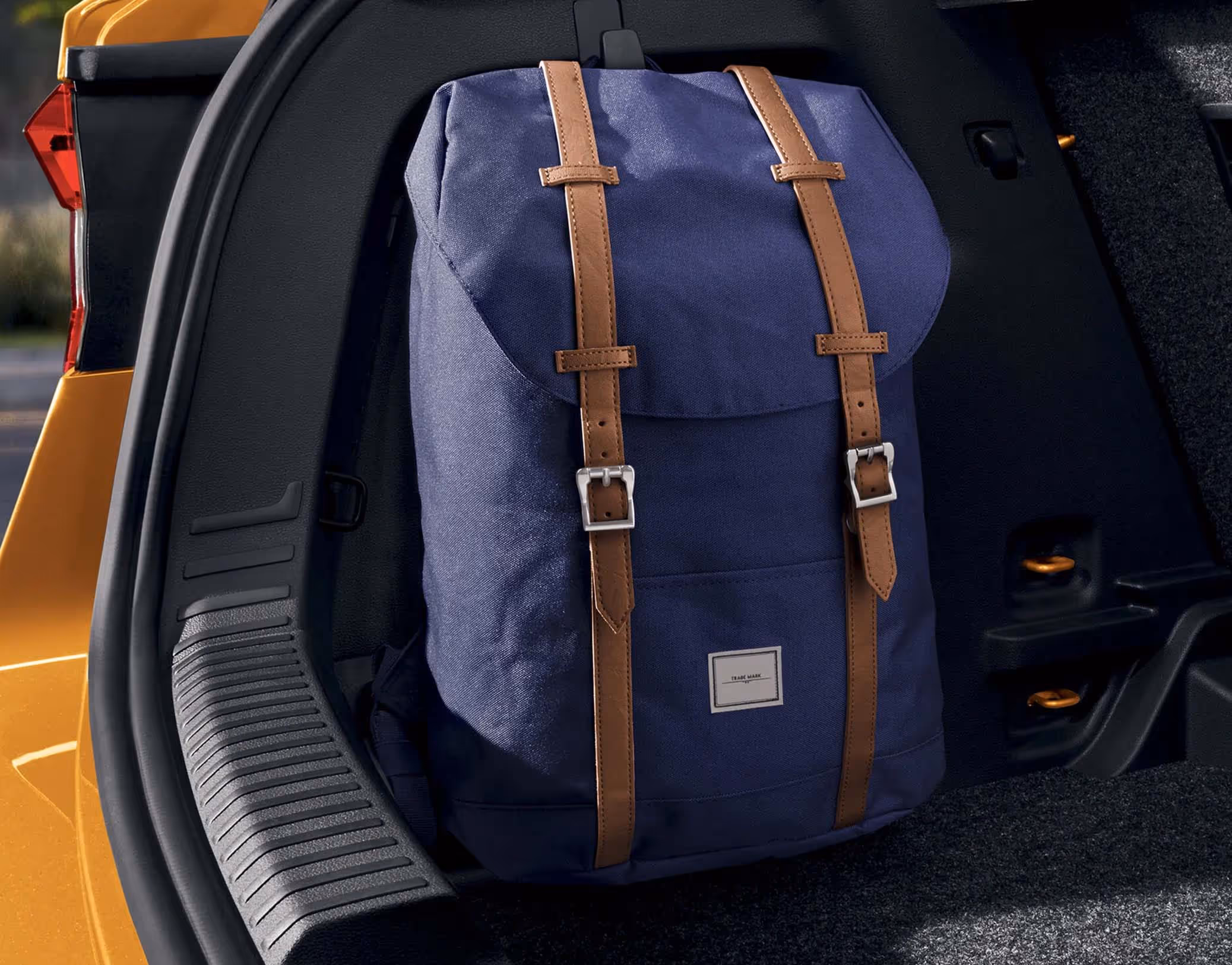 Ein blauer Rucksack mit braunen Riemen hängt an einem robusten Taschenhaken an der Seitenwand des Kofferraums im Škoda Fabia. Diese Vorrichtung fixiert das Gepäckstück sicher in aufrechter Position und verhindert ein Umfallen. Links begrenzen die schwarze Ladekante und die Karosserie in der Lackierung Phoenix-Orange Metallic den Bildausschnitt.