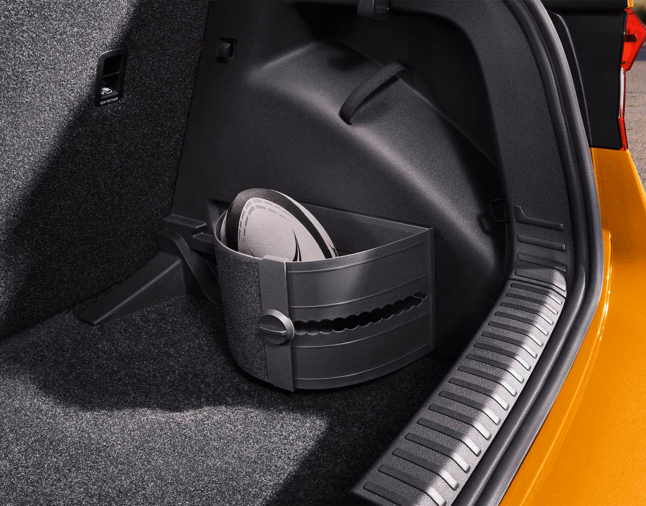 In der seitlichen Nische des Kofferraums bildet eine flexible, gebogene Kunststoffhalterung ein separates Staufach. Darin ist ein Rugbyball fixiert, der so vor dem Verrutschen im Laderaum des Škoda Fabia gesichert wird. Rechts begrenzen die Ladekante und die Karosserie in Phoenix-Orange Metallic den Bildausschnitt.