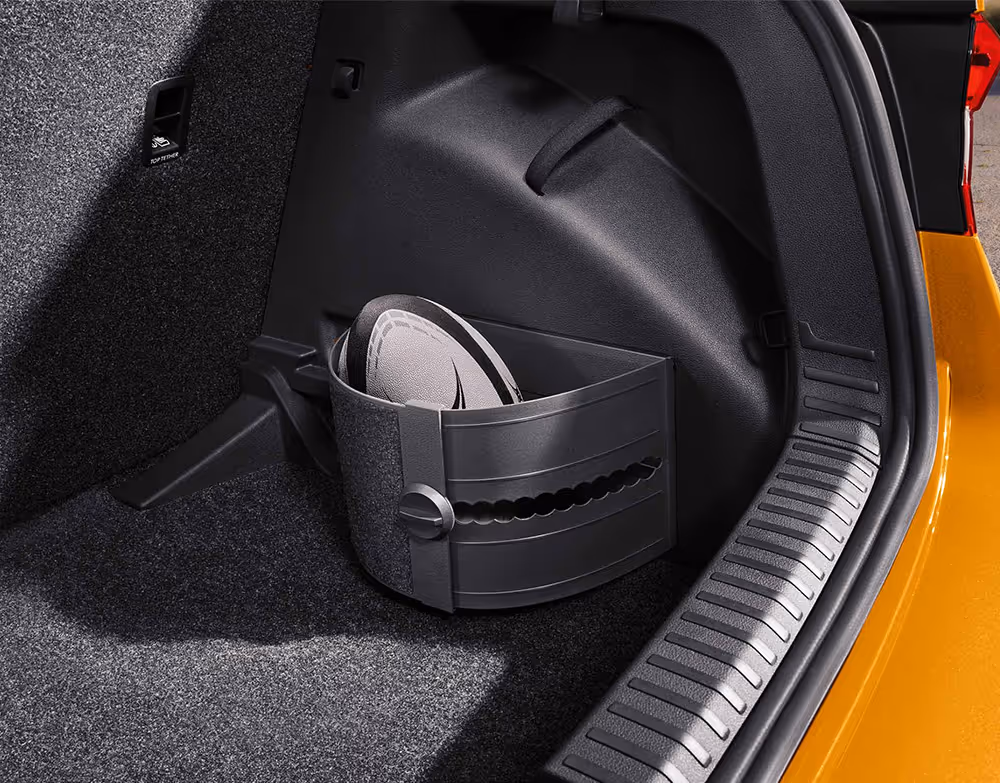 In der seitlichen Nische des Kofferraums bildet eine flexible, gebogene Kunststoffhalterung ein separates Staufach. Darin ist ein Rugbyball fixiert, der so vor dem Verrutschen im Laderaum des Škoda Fabia gesichert wird. Rechts begrenzen die Ladekante und die Karosserie in Phoenix-Orange Metallic den Bildausschnitt.