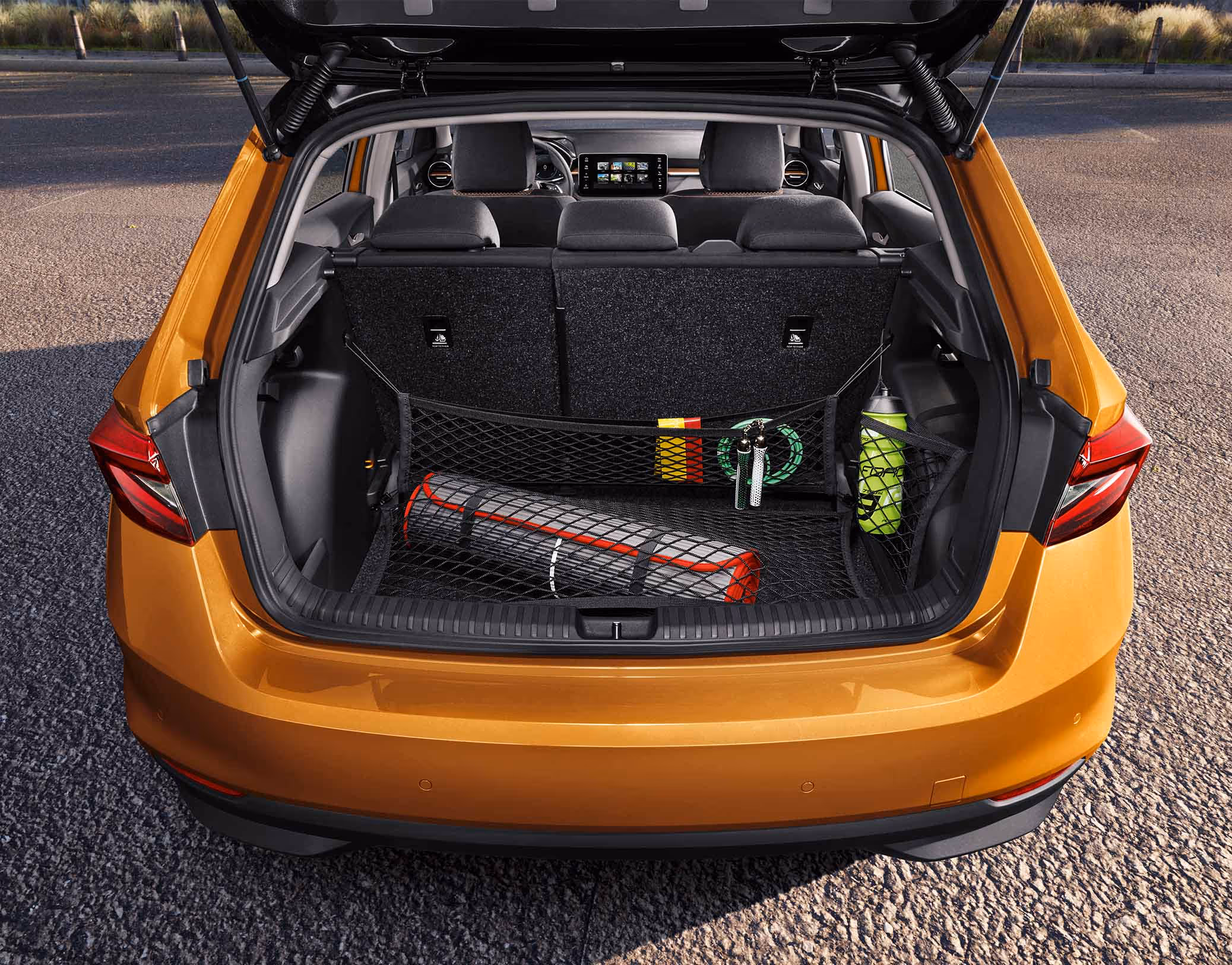 Eine Heckansicht zeigt den geöffneten Kofferraum eines Škoda Fabia in der Lackierung Phoenix-Orange Metallic. Das Netzprogramm sichert diverse Gegenstände wie eine Isomatte und eine Trinkflasche vor der aufrechten Rücksitzbank. Im vorderen Bereich sind die Kopfstützen der Vordersitze sowie das zentrale Infotainment-Display zu erkennen.
