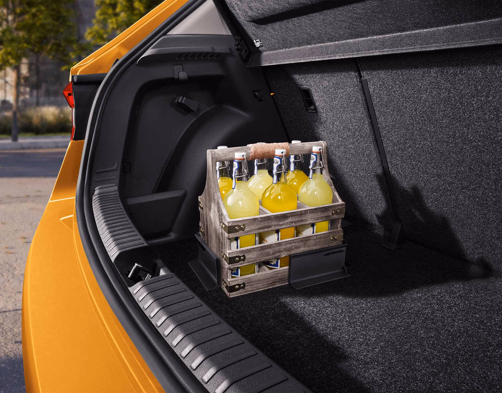 Ein hölzerner Flaschenträger steht auf dem Kofferraumboden des Škoda Fabia und wird durch zwei flexible Cargo-Elemente sicher an seinem Platz gehalten. Die L-förmigen Kunststoffwinkel haften mittels Klettverschluss am Boden des Laderaums und verhindern ein Verrutschen der Ladung während der Fahrt. Links begrenzen die schwarze Ladekante und die Karosserie in Phoenix-Orange Metallic den Bildausschnitt.