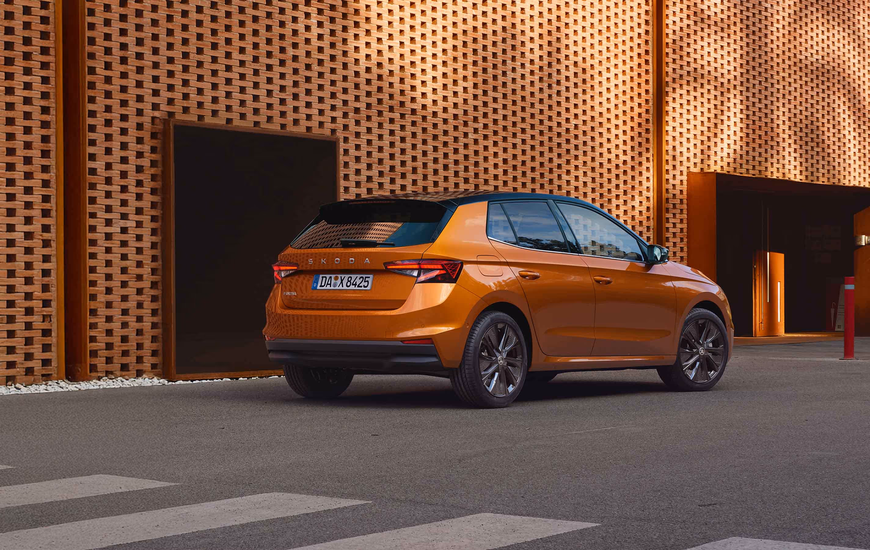 Eine Heckansicht zeigt den Škoda Fabia in der Lackierung Phoenix-Orange Metallic, der mit dem schwarzen Dach des Colour Concept ausgestattet ist. Das Fahrzeug parkt vor einem modernen Gebäude mit einer auffälligen, durchbrochenen Ziegelfassade. Die dunklen Leichtmetallfelgen mit Aeroblenden runden den Auftritt ab.
